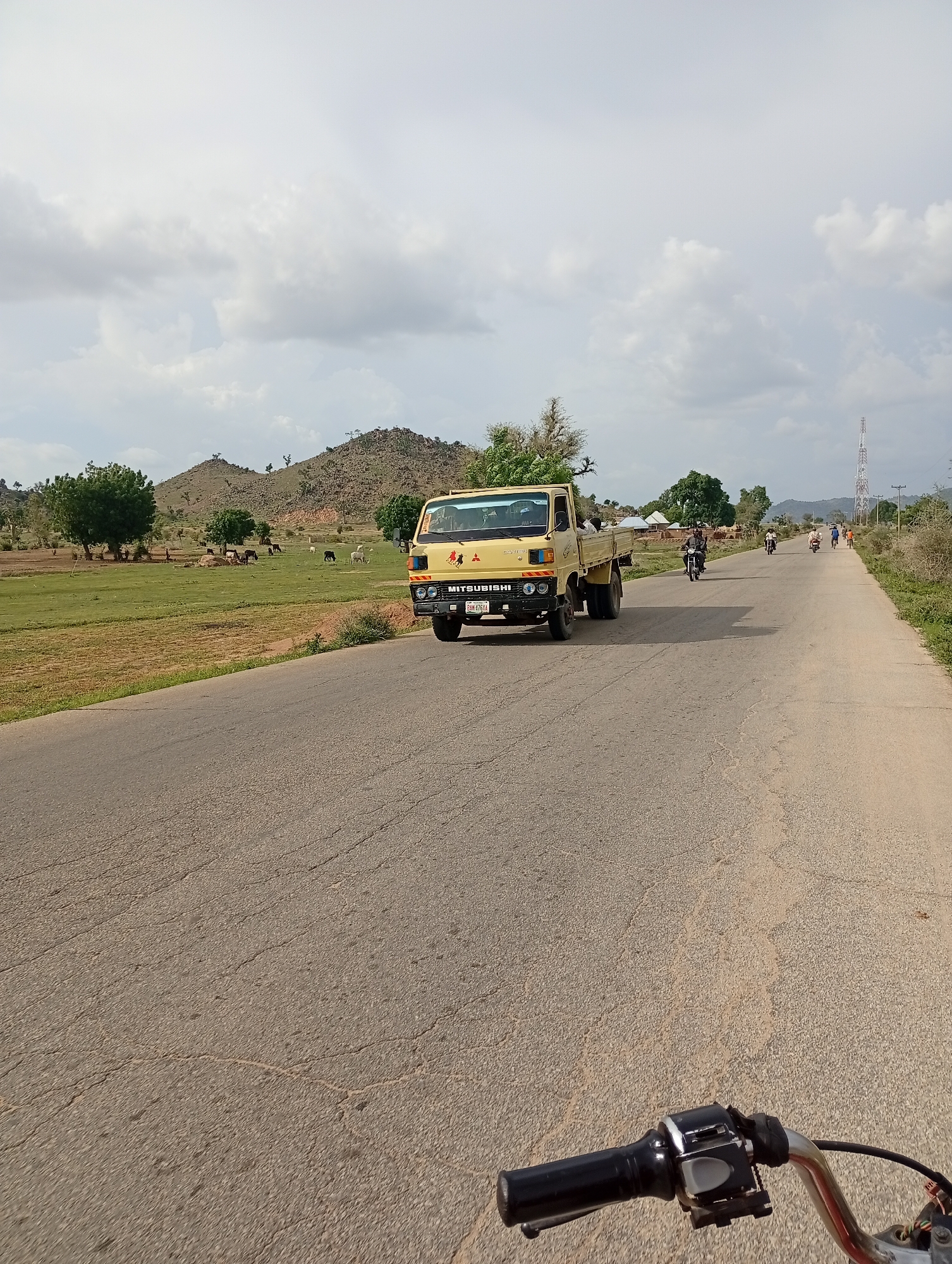 The image shows a road in Shuwa, Nigeria, on a partly cloudy day. A yellow Mitsubishi truck is driving on the right side of the road, with several motorcycles and a few people in the distance. The landscape features rolling hills, green fields, and scattered trees, with a cell tower visible on the horizon. In the foreground, the handlebars of a motorcycle suggest the photo was taken from its perspective. The road is paved and shows signs of wear and tear.