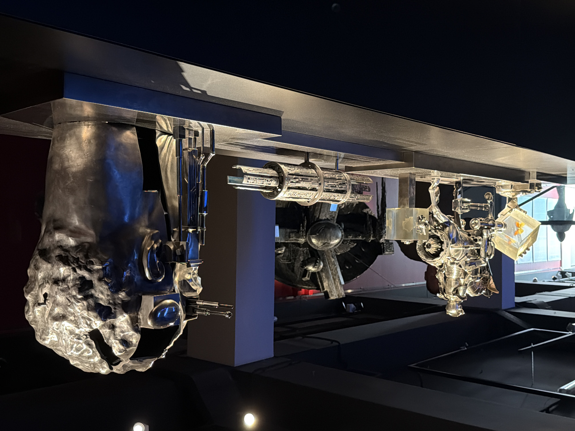 The image displays a museum exhibit featuring several metallic sculptures. In the foreground, a glossy black display counter showcases these artworks. The central sculpture is a silver, segmented cylindrical object supported by stands. To the left, a more intricate sculpture depicts a knight in armor riding a fantastical creature, with a clear acrylic box containing an orange object to its left. Further left, a silhouette of a person is visible near a window. To the right of the cylindrical sculpture is a metallic structure with slender rods. The largest sculpture, dominating the right side of the frame, is a massive, metallic, abstracted head, open at the top, revealing a complex inner mechanism with glass and metal components. The background is dimly lit, with hints of blue and red lighting, and the ceiling features track lighting. The scene appears to be indoors, likely a gallery or museum.