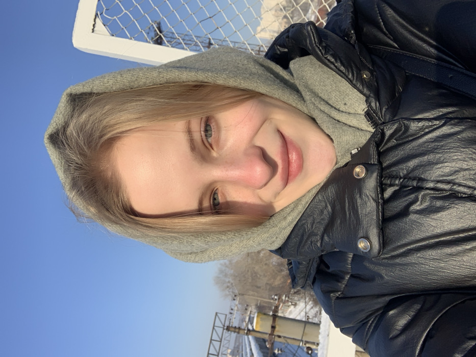 A young female, appearing in her twenties with blonde hair, blue-green eyes, and a fair complexion, smiles directly at the camera. She wears a grey hooded garment around her head and a black, shiny, puffy winter jacket with silver snap buttons on the collar. The image is a brightly lit outdoor selfie taken under a clear blue sky. In the background, to the left, railway infrastructure is visible, including a metal gantry and a tall cylindrical concrete pillar with a yellow band at its upper section, above snow-covered ground and bare trees. To the right, a white metal lattice fence or railing is present. This environment is consistent with the Darnytskyi Bridge (Railway-Automobile Bridge) in Kyiv, Ukraine, specifically on an elevated section overlooking the railway lines.