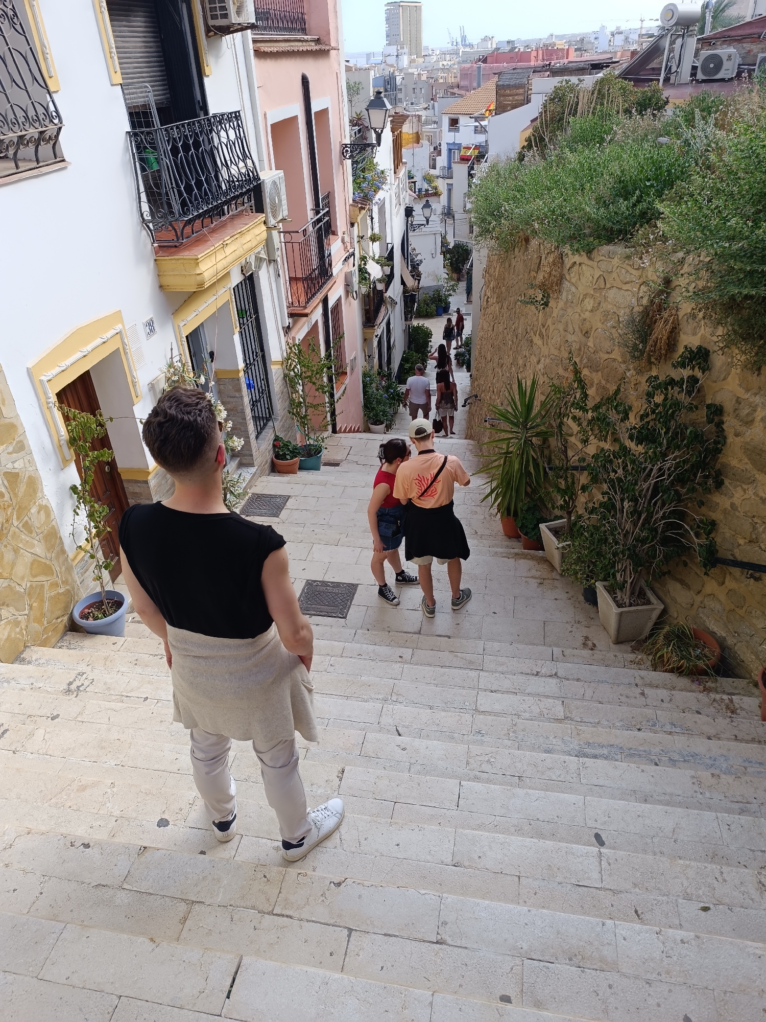 In an outdoor, uphill perspective shot, a young man with short, dark hair and a black t-shirt stands with his back to the camera, facing down a stone staircase lined with buildings. He has a light-colored sweater draped around his shoulders and is wearing light-colored pants and white sneakers. To his left, a white building with yellow trim and an ornate black balcony is visible. To his right, a rough stone wall covered in greenery rises. Further down the stairs, two other individuals are walking away from the camera: a person in a red top and denim shorts, and another person in an orange t-shirt, black shorts, and a white baseball cap. The staircase descends through a narrow street with colorful buildings and potted plants. In the background, a cityscape with various buildings, some tall, stretches towards the horizon. The lighting suggests it is daytime, possibly late afternoon, with soft shadows.