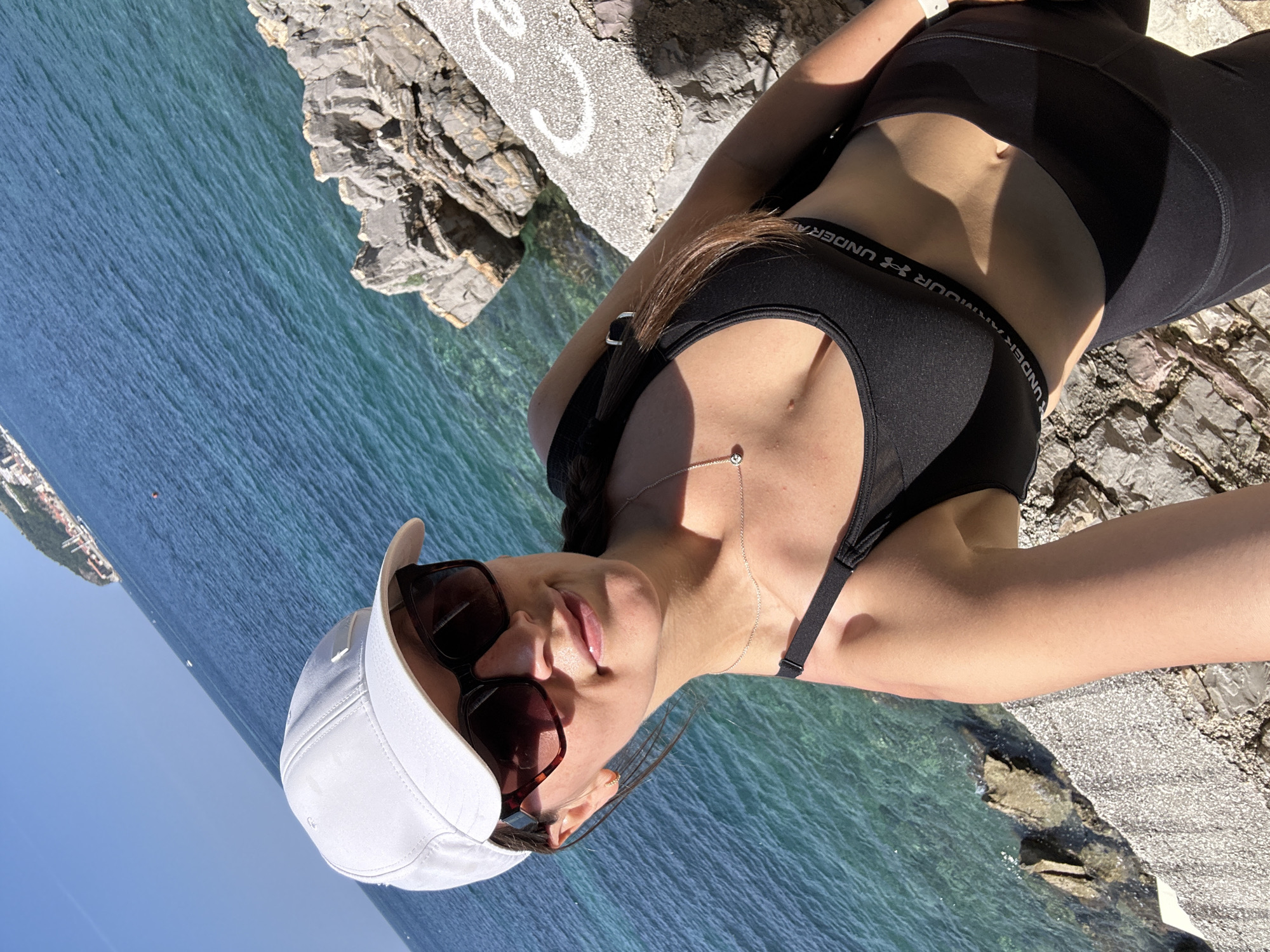 A woman wearing a white baseball cap, dark sunglasses, and a black sports bra takes a selfie outdoors. She is positioned on a rocky embankment with the blue sea and a distant coastline visible in the background. The woman has brown hair that is partially braided and she is wearing a silver necklace with a small pendant. The scene is brightly lit by sunlight.