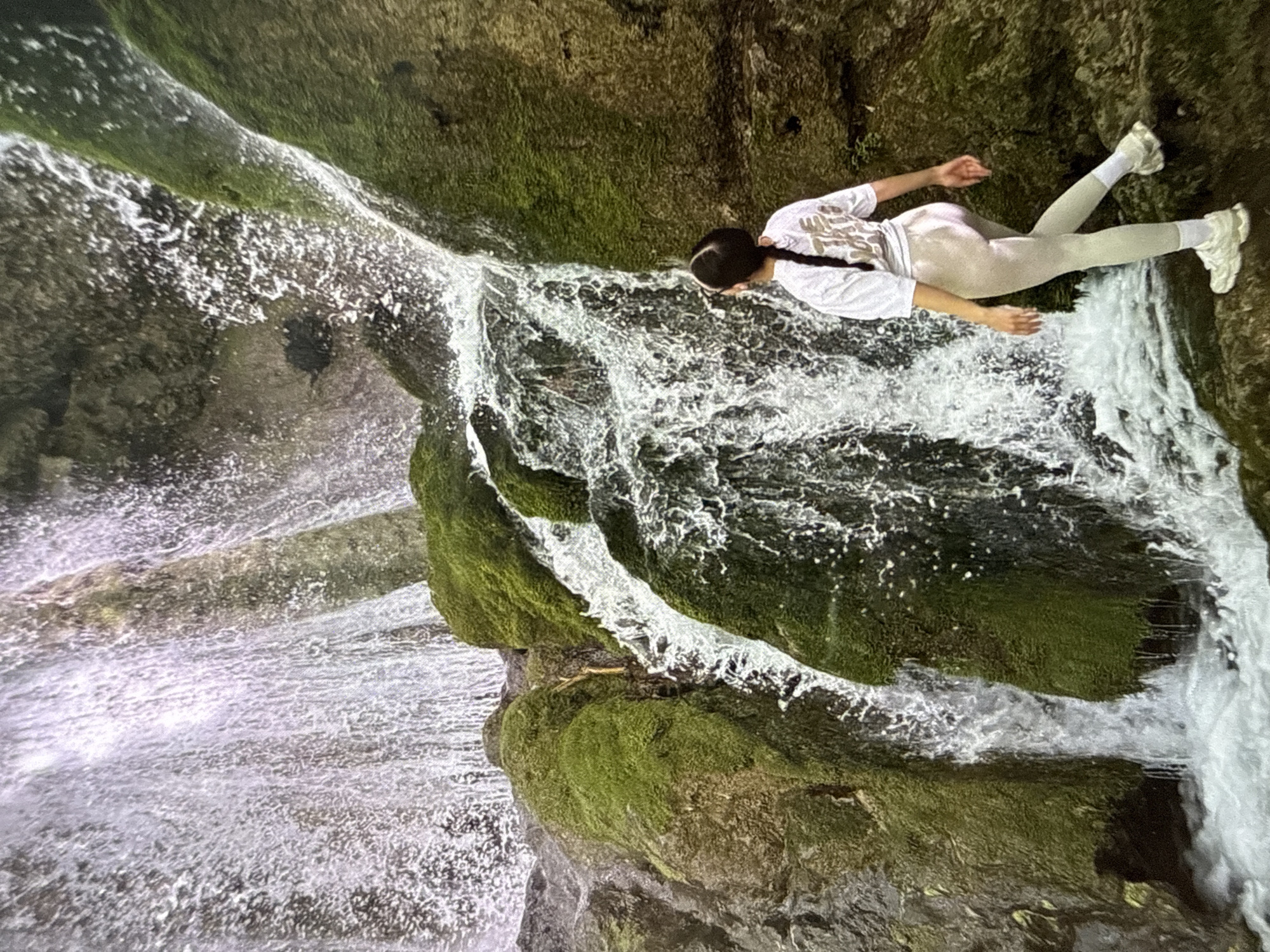 A woman is lying on a rock covered with moss next to a waterfall. She is wearing a white t-shirt, light-colored leggings, and white sneakers. Her hair is in a braid. The waterfall has multiple tiers of water cascading over rocks covered in green moss. The location is in the city of Ivaylovgrad, Bulgaria.