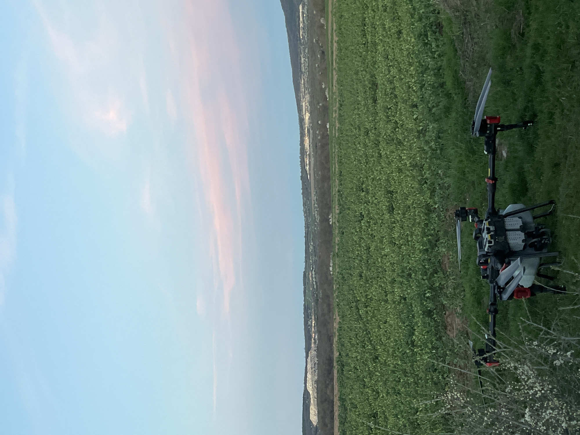 A color, landscape-oriented image depicts a rural scene with a drone in the foreground. The location is Varbeshnitsa, Bulgaria. To the left is a light blue sky with hints of pink clouds. The center portion shows a strip of land separating a field of green vegetation from a settlement of white buildings and greenery. In the foreground to the right, there's a multi-rotor drone equipped with a tank, likely for agricultural spraying. Some branches with white flowers obscure a part of the drone.