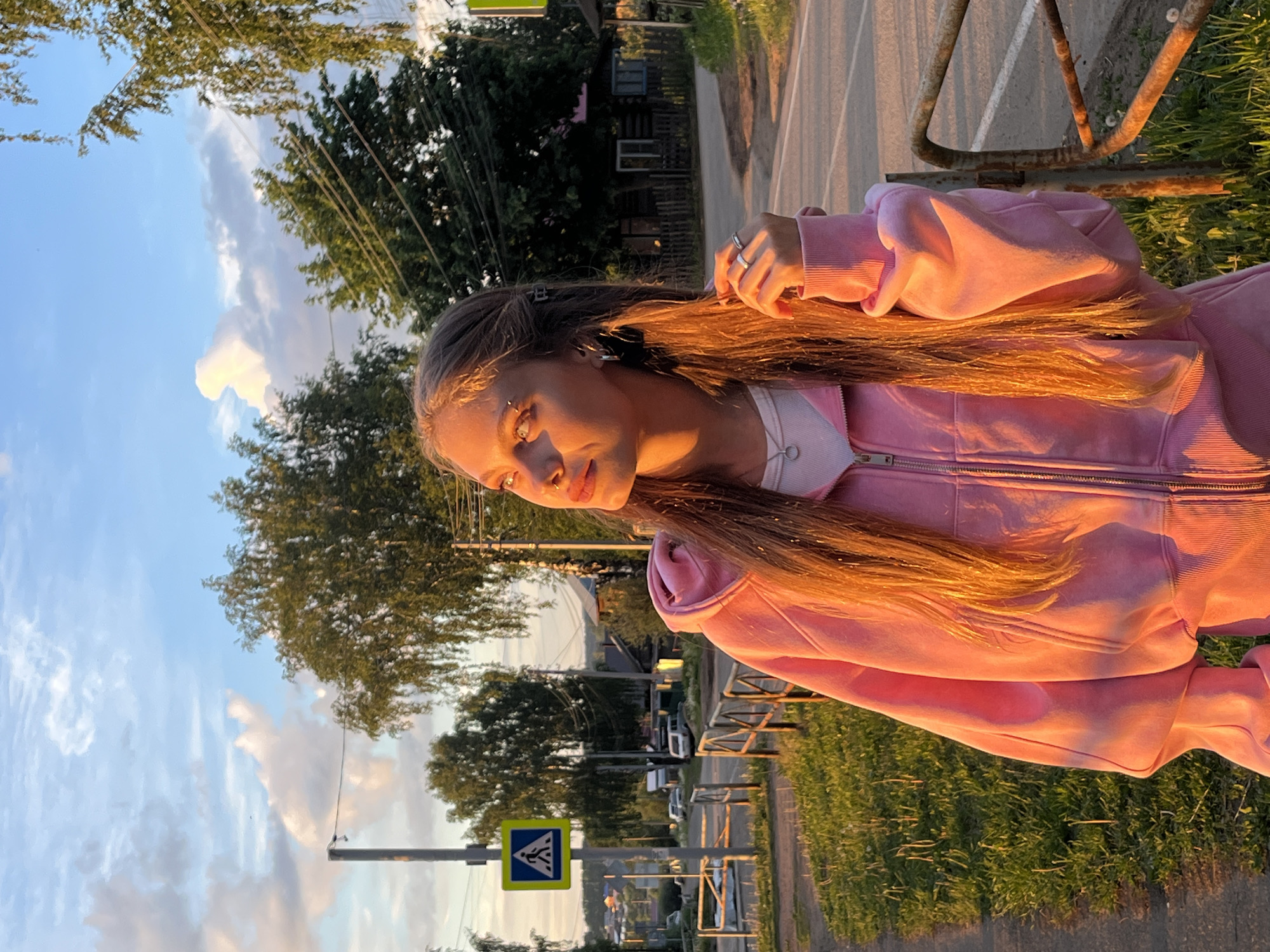 A young woman stands on the side of a street in Фоки, Russia. She wears a pink hoodie and runs her hand through her long brown hair. Behind her is a street with trees, grass, and a crosswalk sign. The sky is blue with scattered clouds.