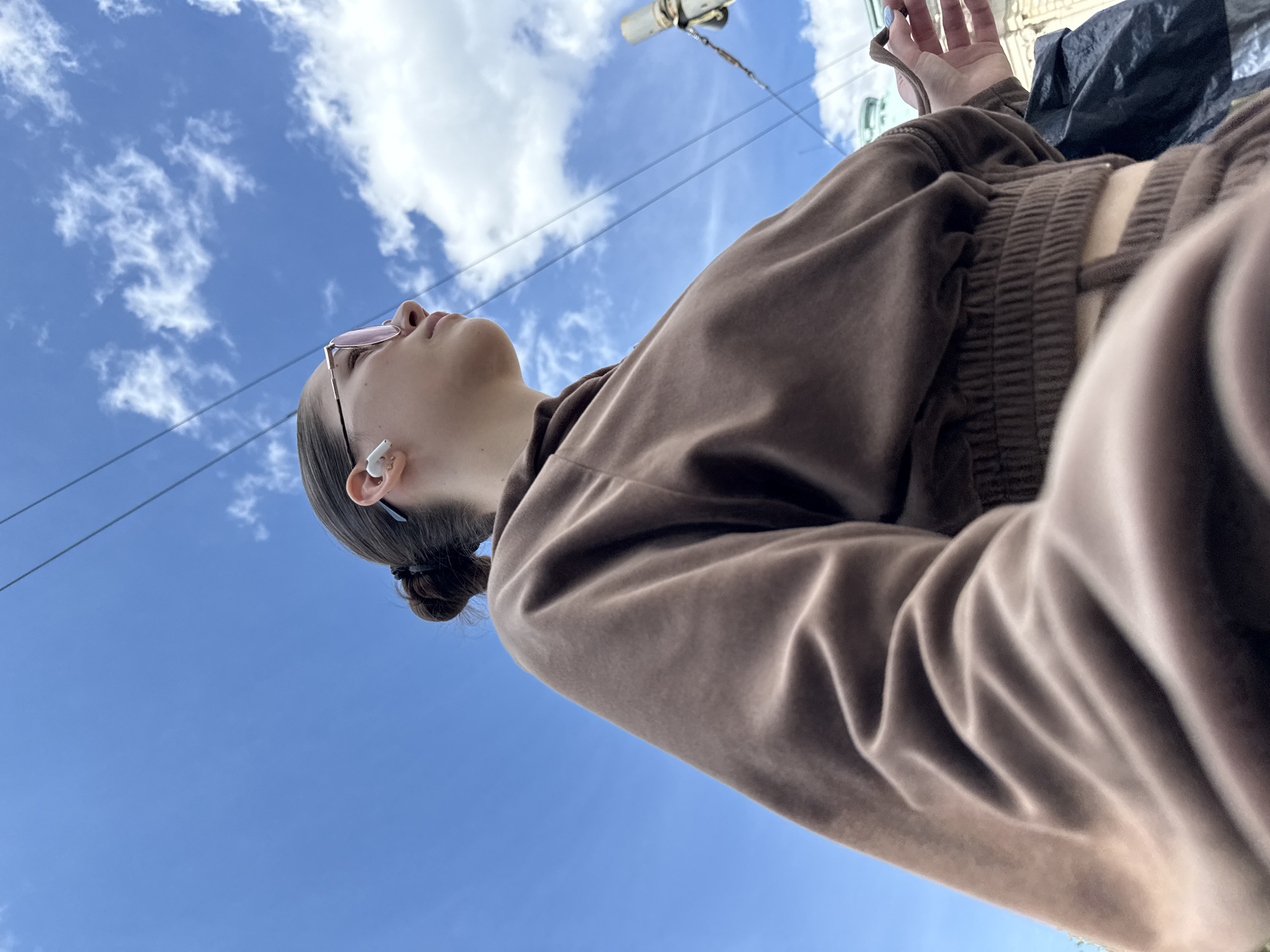 A low-angle shot depicts a young woman with light skin and brown hair pulled back into a bun. She is wearing pink-tinted glasses and a white wireless earbud in her left ear. The woman is dressed in a brown, possibly velour, crop top and matching sweatpants or shorts. The image appears to have been taken outdoors, with a blue sky and scattered clouds visible in the background.
The photograph might have been taken in Sofia, Bulgaria. The presence of power lines and urban structures in the background hints that the location is in a populated area, as does a building.