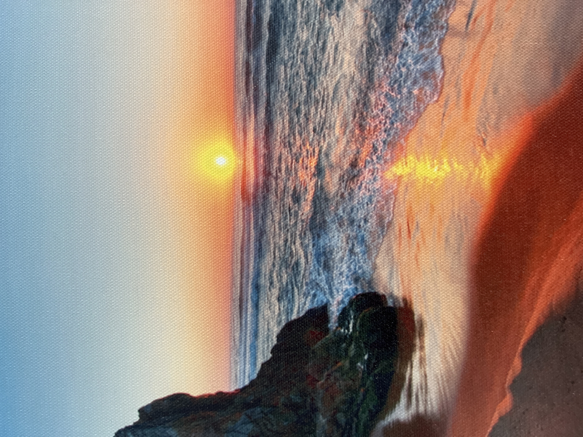 The image depicts a scenic sunset over a beach in Eretria Municipal Unit, Greece. The sky transitions from a clear blue at the top to hues of orange and yellow near the horizon, where the sun is visible as a bright, small disc. The sea is calm, with gentle waves washing onto the sandy shore, reflecting the warm colors of the sunset. On the left side of the image, a rocky outcrop with patches of greenery juts out into the sea. The wet sand in the foreground also mirrors the vibrant sunset colors, creating a striking visual contrast. There are no people or animals visible in the scene. The image appears to be a photograph or a painting on canvas, as indicated by the textured surface.