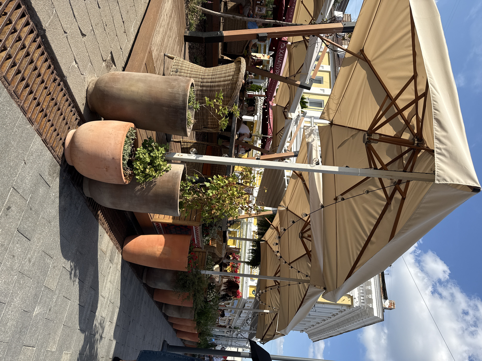 The image depicts an outdoor seating area of a cafe or restaurant with tables and chairs arranged under large beige umbrellas. In the foreground, several large terracotta pots contain plants, with one pot to the left appearing to be made of a lighter, grayish material. A metal grate for drainage runs along the edge of the paved walkway. The seating area itself is on a wooden deck. In the background, a yellow building with white architectural details is visible, suggesting an urban setting. Several people are seated at tables, some interacting, while others appear to be dining or conversing. The overall impression is of a sunny day with ample shade provided by the umbrellas. The cafe appears to be "Хачапури," as indicated by signage visible on the red awnings in the background. This establishment is located in Nizhny Novgorod, Russia.
