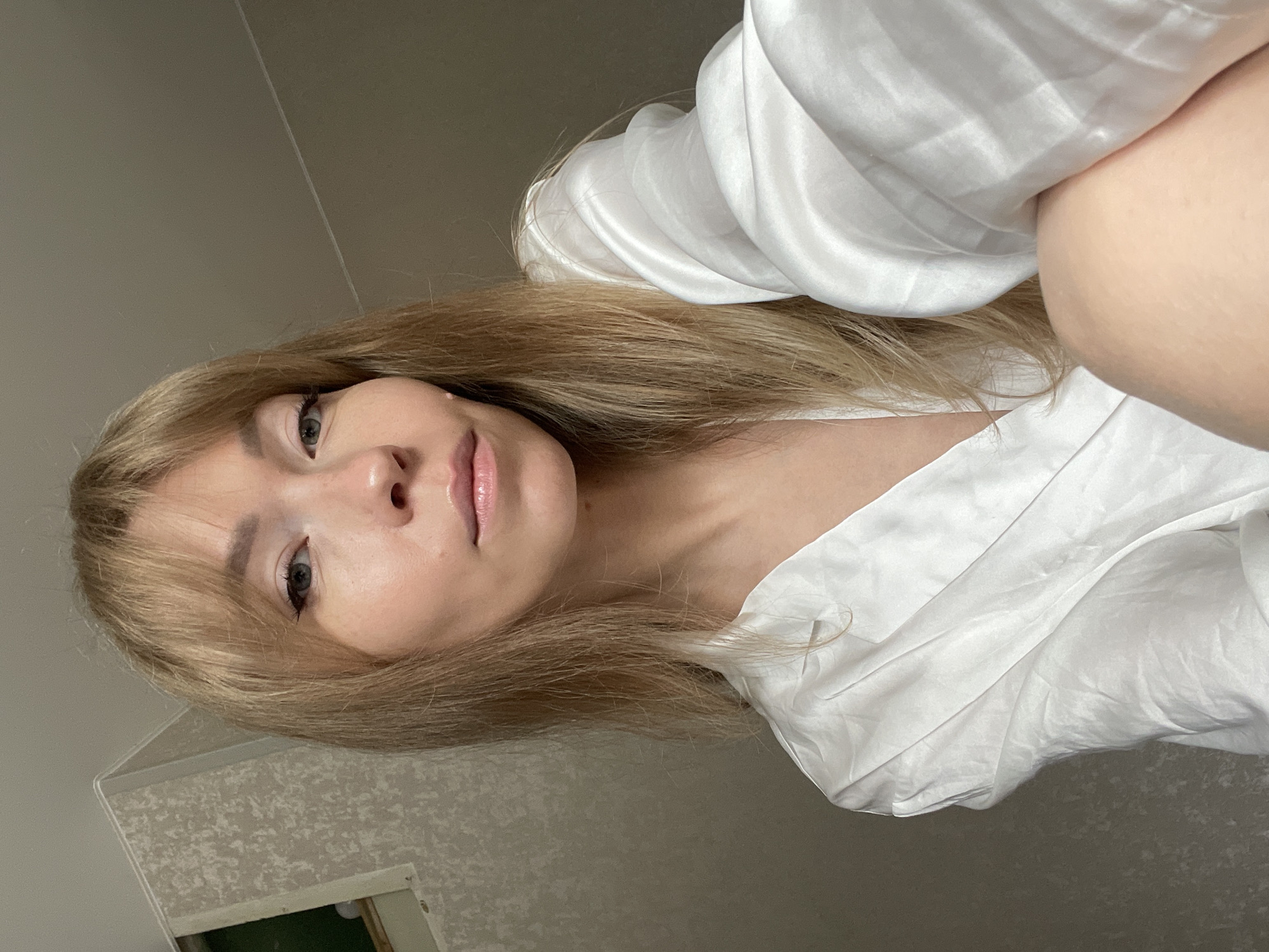 A woman with blonde hair and bangs, wearing a white satin robe, is shown in a close-up selfie. She has light skin, blue eyes with eyeliner, and her lips are slightly parted and glossy. Her hair is long and straight, falling over her shoulders and the front of her robe. The background appears to be an indoor setting, possibly a bedroom, with textured wallpaper on the left and a plain, light-colored wall on the right. The lighting is soft, casting a gentle glow on her face. The image quality is good, with clear focus on the woman. There is no discernible location in the city of Perm, Russia, or any context to suggest it.