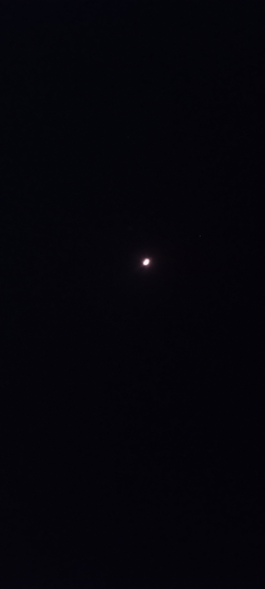 The image is a nighttime photograph of the sky. A crescent moon is visible, emitting a soft glow. To the right of the moon, a small, faint star can be seen. The background is entirely dark, suggesting a clear night sky. There are no visible landmarks, buildings, or any indication of the city of Adai, Spain, within the frame. The photograph captures a celestial scene with no human presence or activity.