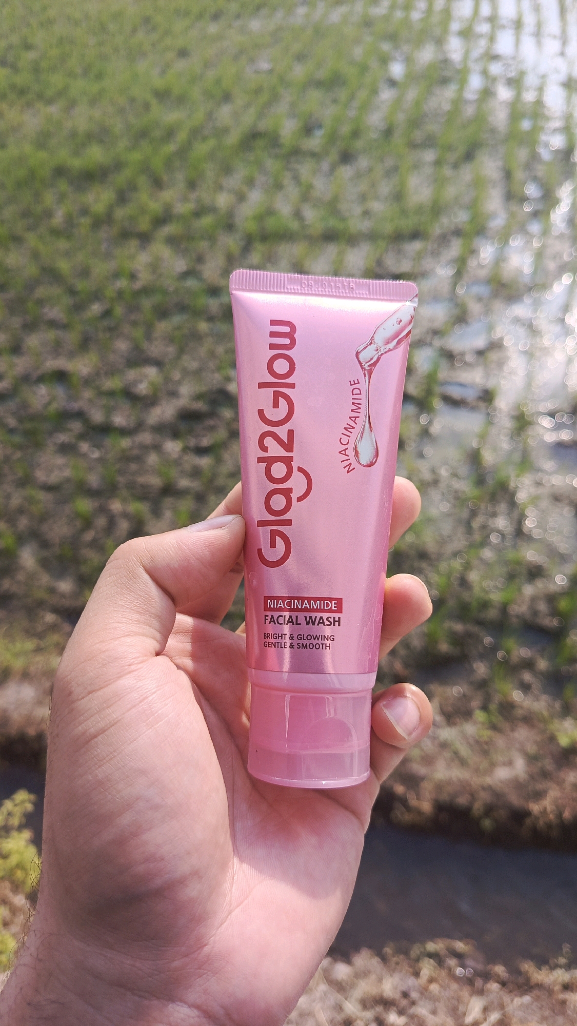 An outdoor, eye-level, medium shot captures a person's hand holding a pink tube of "Glad2Glow" facial wash. The product features red lettering and a graphic of a dropper. The background is a blur of green rice paddies and a water channel, suggesting a rural setting, possibly in Babadan, Indonesia. The lighting is natural and bright, with sunlight glinting off the water in the background.