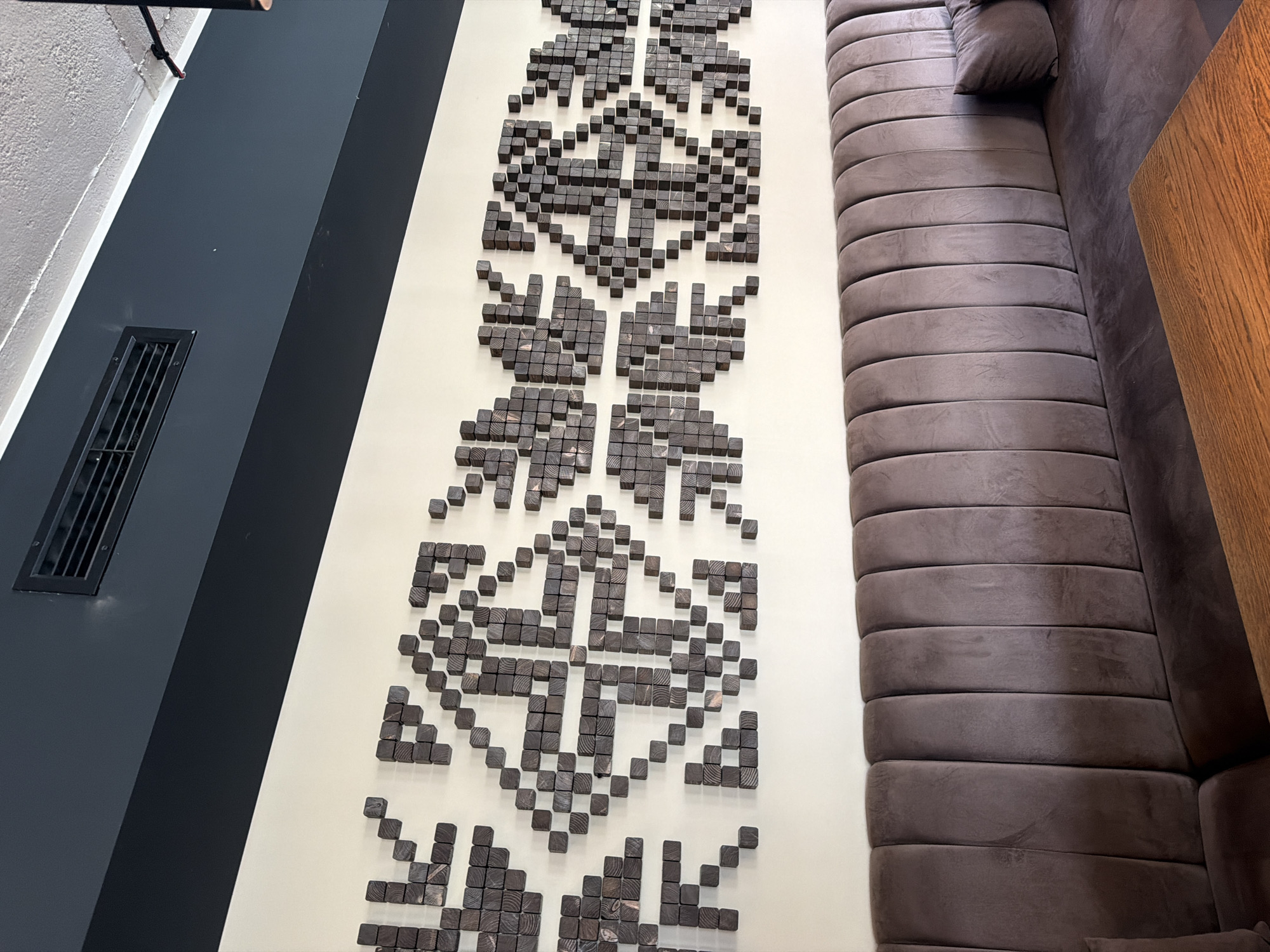 The image depicts the interior of a commercial establishment, likely a cafe or restaurant, located in the city of Sofia, Bulgaria. The primary focus is a long, light-colored wall adorned with an extensive horizontal art installation composed of numerous small, dark wooden blocks. These blocks are arranged in a repeating, pixelated, geometric pattern that spans the length of the wall, resembling traditional folk motifs.
Below the wall art, a continuous, dark brown or taupe upholstered bench extends across the lower portion of the frame. The bench features a vertically channeled backrest made of a velvet-like fabric, with a flat seat cushion. A dark throw pillow is visible on the right end of the bench. A wooden table with a prominent grain and a warm brown hue is partially visible in the bottom right foreground.
Above the light-colored wall, a dark charcoal or slate-grey section of a wall or beam runs horizontally, separating the lower wall from the ceiling. A rectangular black air conditioning vent with horizontal louvers is installed within this dark section on the left side. The ceiling above is a lighter, textured surface, possibly concrete or painted plaster, with a thin white trim line demarcating it from the dark wall section. A small white speck is visible on the dark wall section to the right of the vent. A black fixture, possibly a light or speaker, is partially visible extending from the ceiling on the far right. No individuals are present in the scene.