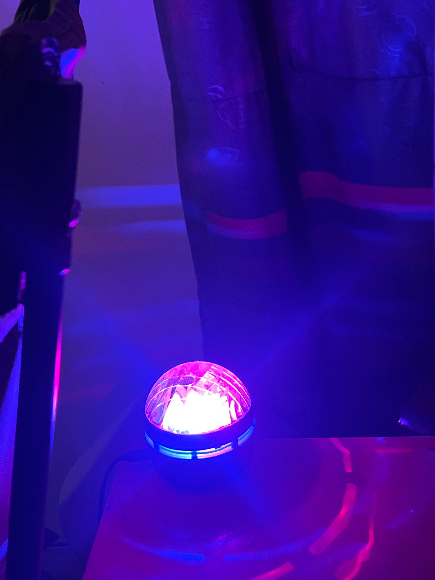 The image shows a disco ball illuminated with pink and blue lights, sitting on a flat surface. The surface is reflective, showing streaks of light. In the background, a dark, textured curtain or fabric is visible, also lit by the colored lights. The overall atmosphere is dark and moody, typical of a party or club setting. There is no visible indication of Sleman, Indonesia.