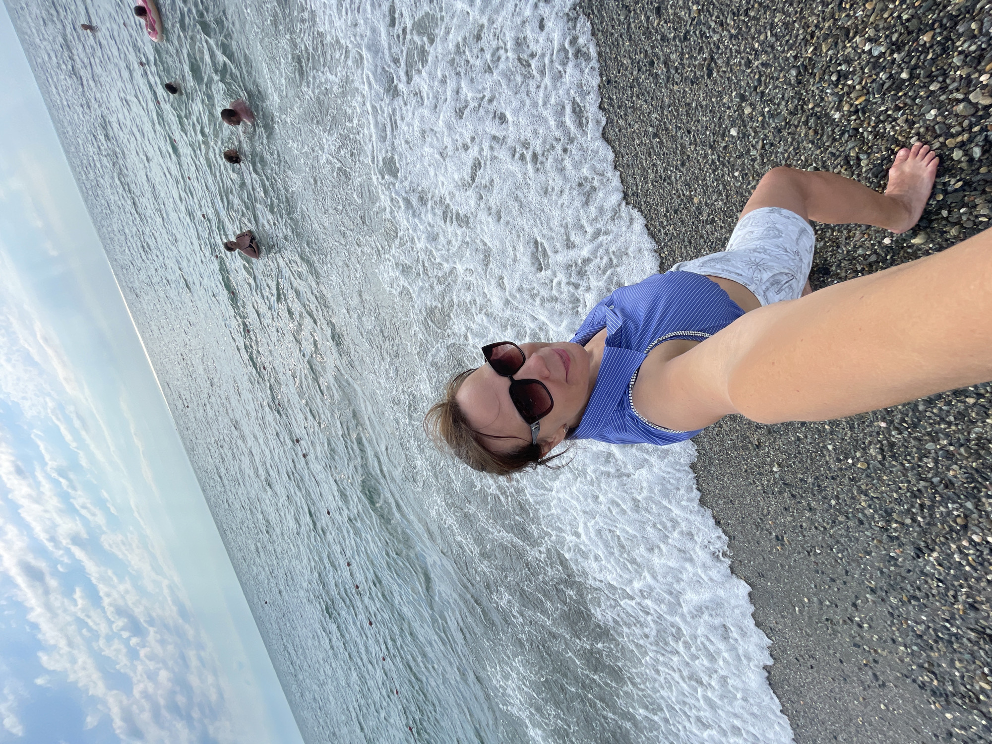 A woman is taking a selfie on a pebble beach in Sochi, Russia. She is wearing a blue and white striped top and white shorts. She has sunglasses on. The sea is calm with small waves breaking on the shore. In the background, several people are swimming in the sea and one person is on an inflatable ring. The sky is cloudy with patches of blue.