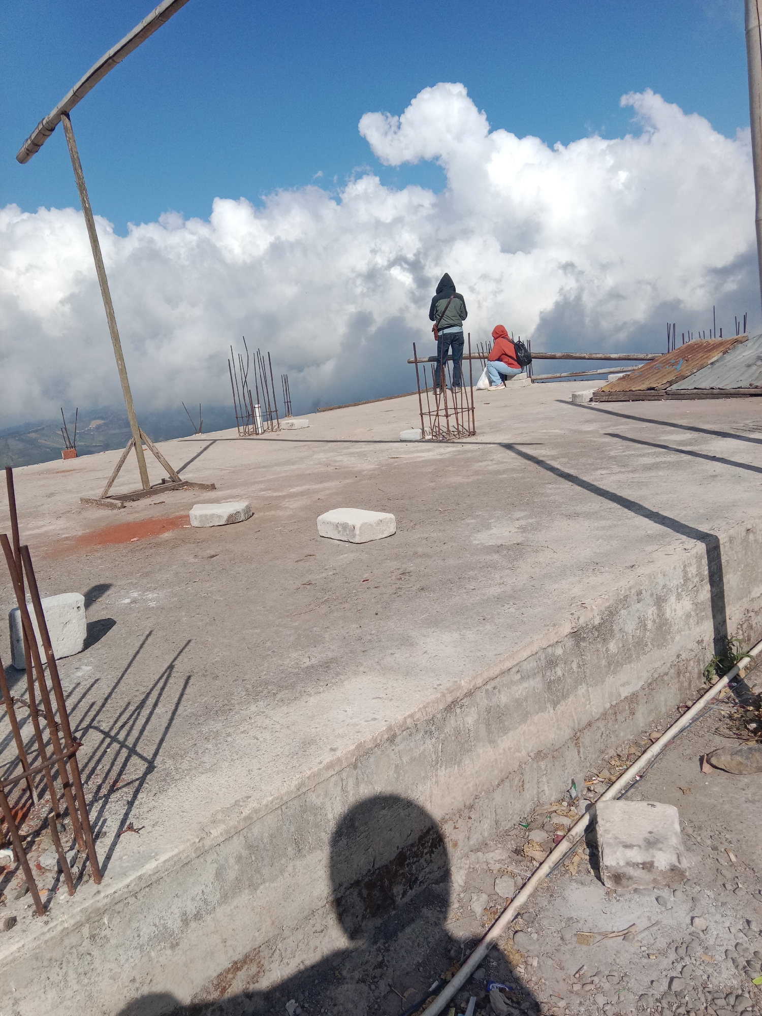 The image depicts a rooftop under construction, likely at a high altitude given the presence of expansive clouds below and a clear blue sky above. Two individuals are present on the rooftop. One person, wearing a dark hooded jacket and jeans, stands facing away from the camera, looking towards the cloudscape. The second individual, wearing a red hooded jacket and jeans, is seated with their back to the camera, also observing the view. The rooftop is made of concrete and shows signs of ongoing work, with rebar protruding from various points and several concrete blocks scattered across the surface. A rusted metal sheet is visible in the background. The scene appears to be in a remote location, possibly Tieng, Indonesia, given the typical terrain and atmospheric conditions described for such areas. The lighting suggests it is daytime, with strong shadows cast by the rebar and the photographer's presence.