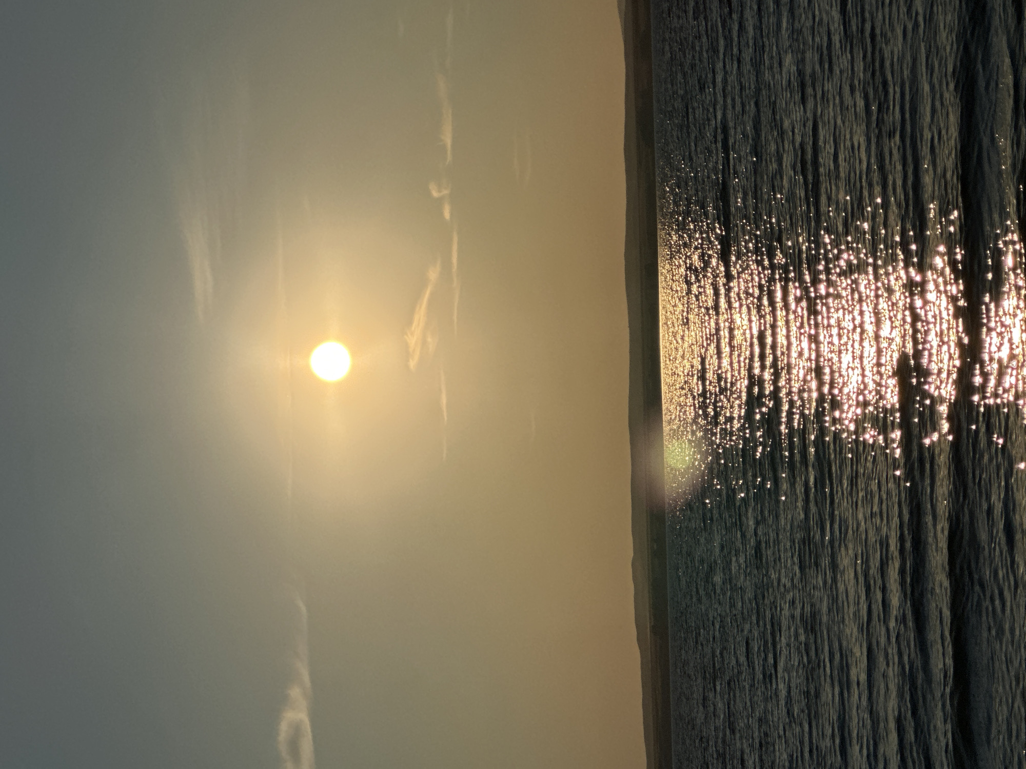 The image depicts a serene sunset over a body of water, likely a reservoir, near the city of Energetik, Russia. The sun, a bright, warm orb, hangs low in the sky, casting a golden glow that reflects shimmeringly on the gentle waves. The water itself is a deep blue, contrasting with the lighter hues of the sky and the distant, silhouetted horizon line of rolling hills. A faint, wispy cloud streaks across the upper left quadrant of the sky, adding a subtle texture to the otherwise clear expanse. The overall atmosphere is one of tranquility and natural beauty, capturing a moment of peaceful transition from day to night.