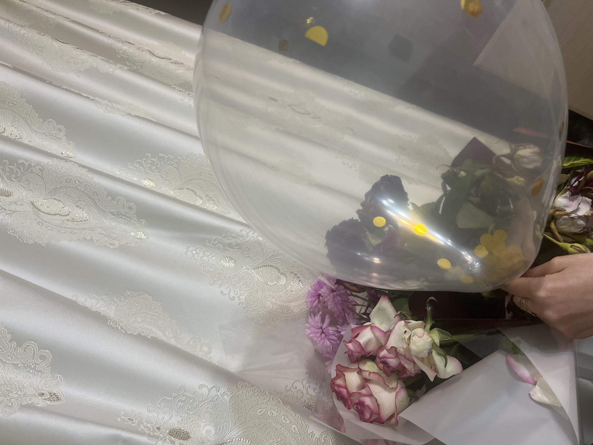 A close-up, angled shot shows a bouquet of white and pink roses and purple mums in front of white, textured curtains. A clear balloon filled with confetti and dark flowers partially obscures the bouquet. A hand wearing two rings is touching the balloon. The scene appears to be indoors, possibly in a domestic setting in Dubăsari town council, Moldova, given the presence of a bouquet and balloon which are common for celebrations.