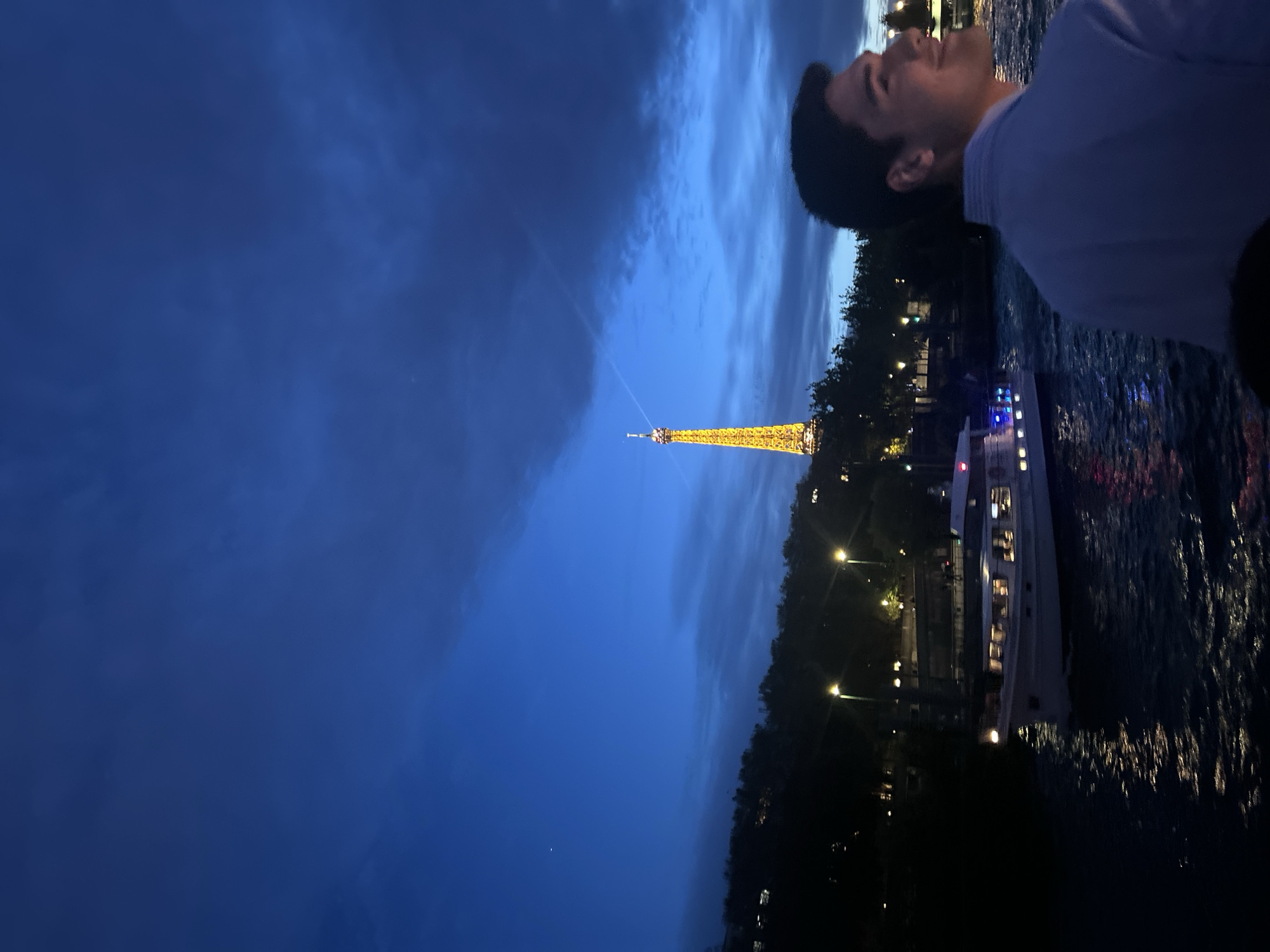 A medium-shot photograph shows a man looking upward toward the Eiffel Tower in Paris, France. The scene is set at night, with the sky a deep blue interspersed with streaks of clouds. The Eiffel Tower is illuminated in gold. Adjacent to the man, a tourist boat is afloat on the river, lit with colorful lights. Trees and streetlights are also visible along the riverbank. The image is rotated 90 degrees clockwise.