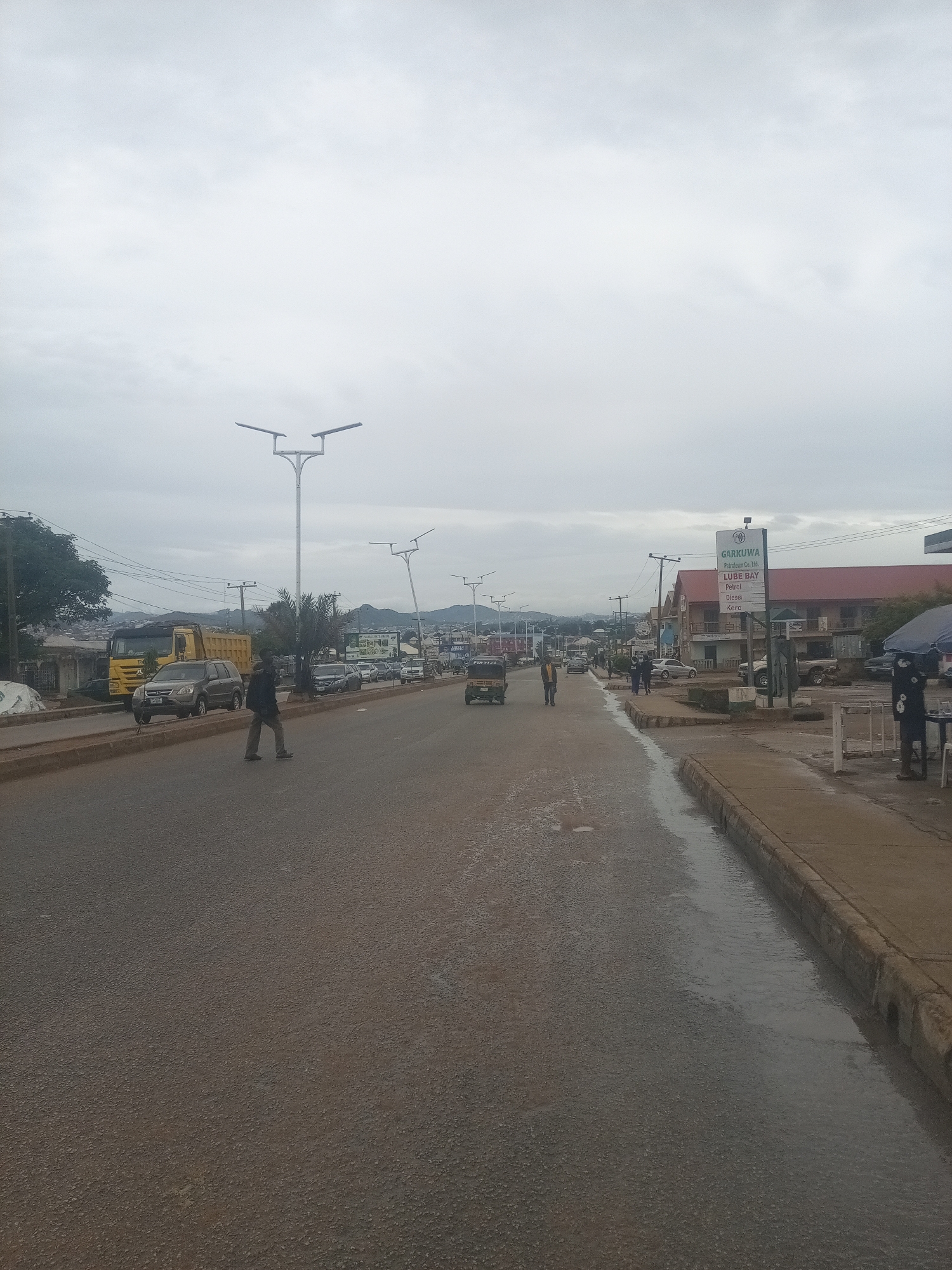 A street scene unfolds under an overcast sky, likely in Rafin Par, Nigeria. The road is wet, with puddles reflecting the muted light and a channel of water running along the curb on the right side of the image. A man is crossing the road in the foreground, walking from left to right. Several vehicles are visible: a yellow dump truck parked on the left, a dark SUV, and other cars and a trishaw further down the street. Lampposts with modern designs line the roadside. On the right side of the street, a building with a red roof features a prominent sign for "GARKUWA Petroleum Co. Ltd. LUBE BAY," advertising Petrol, Diesel, and Kero. Two figures, possibly staff, are standing near an umbrella-covered stall on the sidewalk. In the distance, hills or mountains are visible. The overall impression is of a typical urban street scene, possibly after a rainfall.