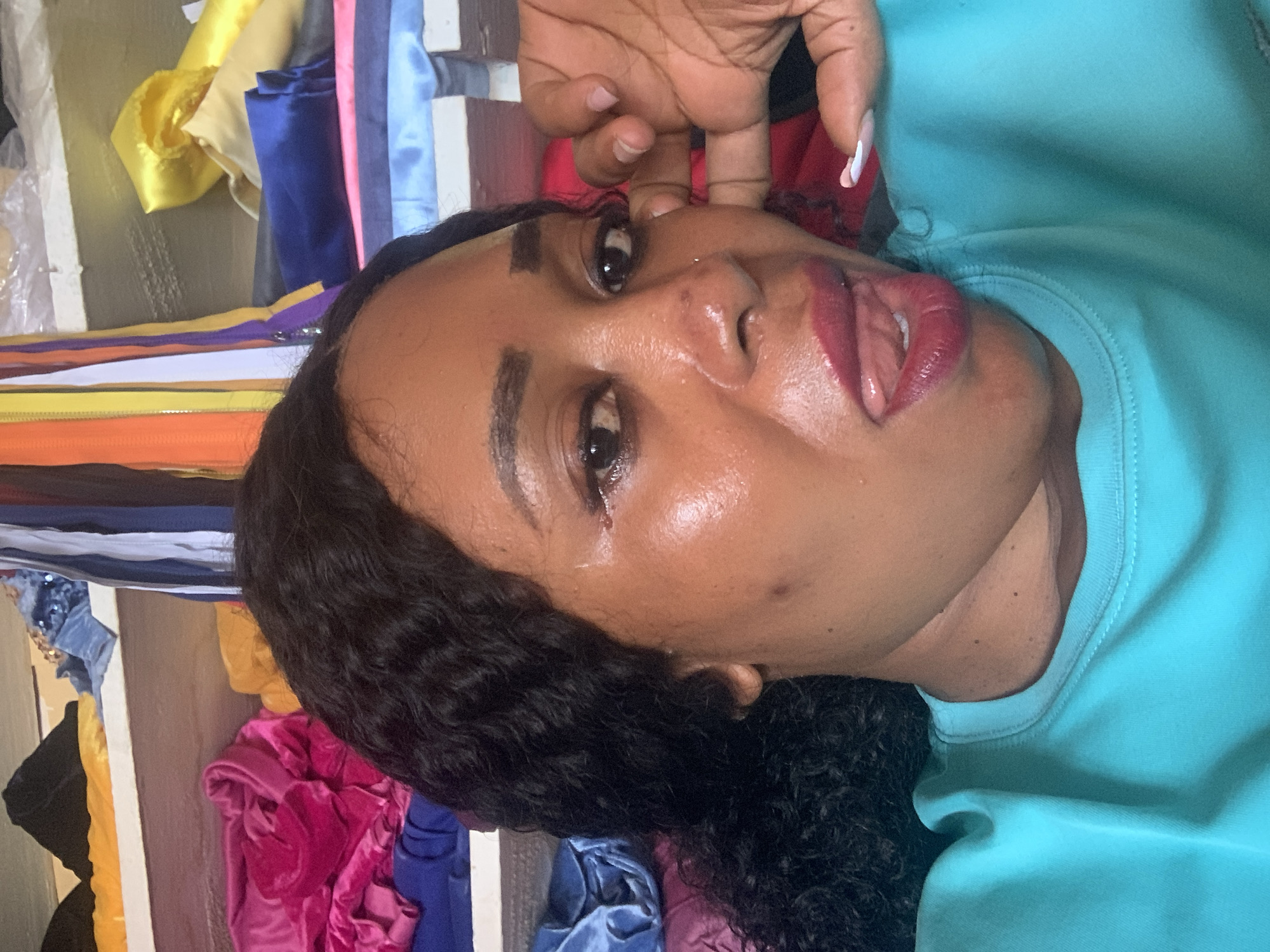 A woman is posing for a selfie. She has brown skin, dark hair, and is wearing a blue top. Her fingers are near her face. Behind her are various fabrics of different colors hanging on a rack. It is not possible to confirm the location.