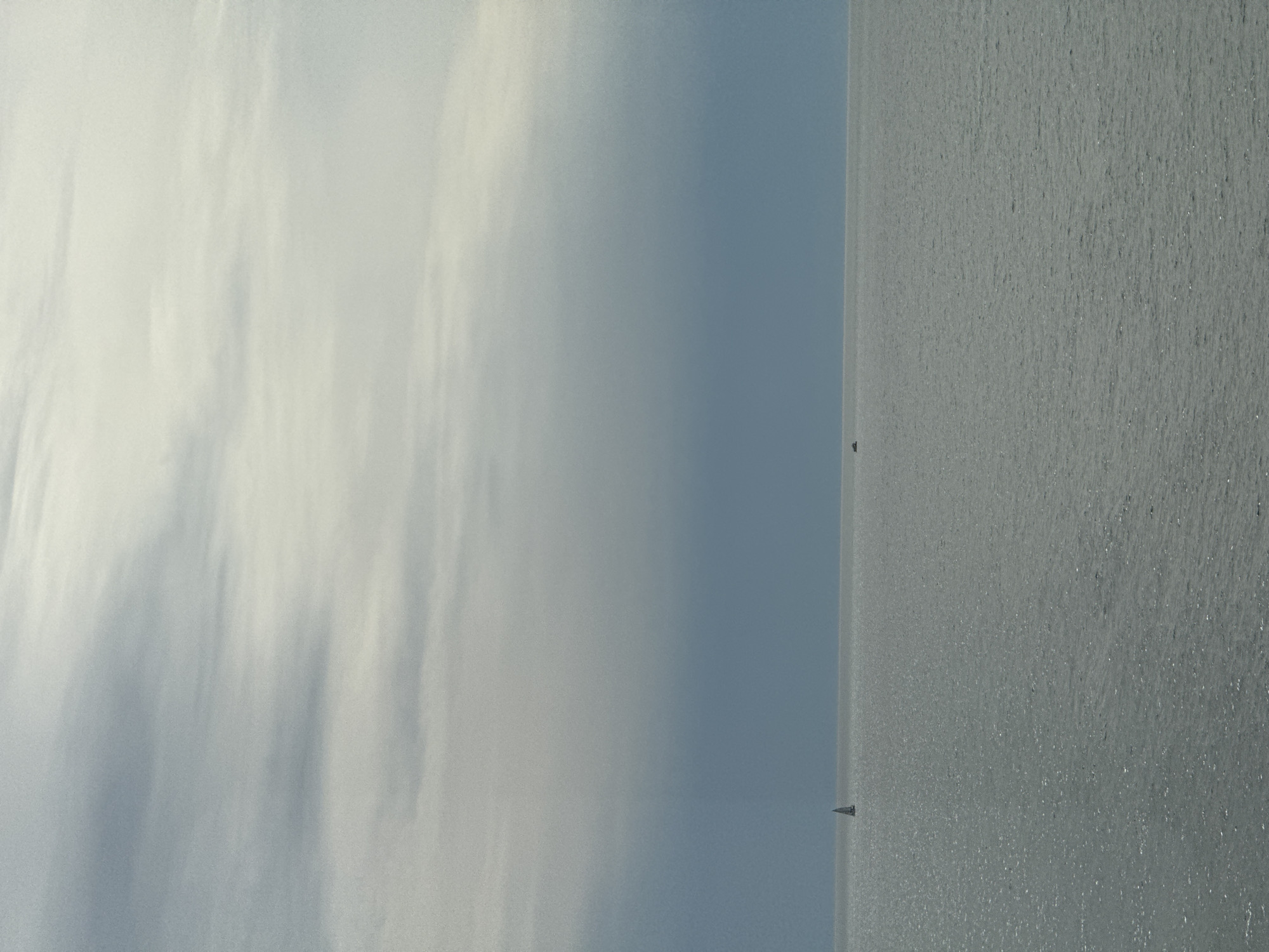 The image depicts a body of water, likely the Black Sea, adjacent to the city of Sochi, Russia. The right side of the image shows the water, with small, bright reflections suggesting sunlight. Two small sailboat silhouettes are visible on the water's horizon. The left side shows the sky with light clouds. The horizon line between the water and sky is straight and distinct. The perspective indicates the photo was taken at eye level, likely from the shore.