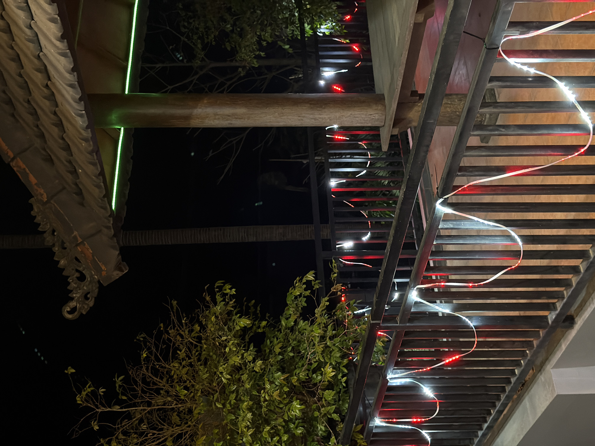 The image shows a nighttime scene on a balcony or deck area. A wooden railing is prominent in the foreground, strung with decorative lights that alternate between red and white. Behind the railing, a covered structure with a tiled roof and a wooden beam is visible, illuminated by a green neon light strip. A tree with green foliage is to the left, and bare branches are visible in the background against the dark night sky. The environment appears to be an outdoor seating or gathering space, likely in a tropical or subtropical location given the foliage. The image does not contain any people or specific actions. The location is identified as Wonoayu, Indonesia.