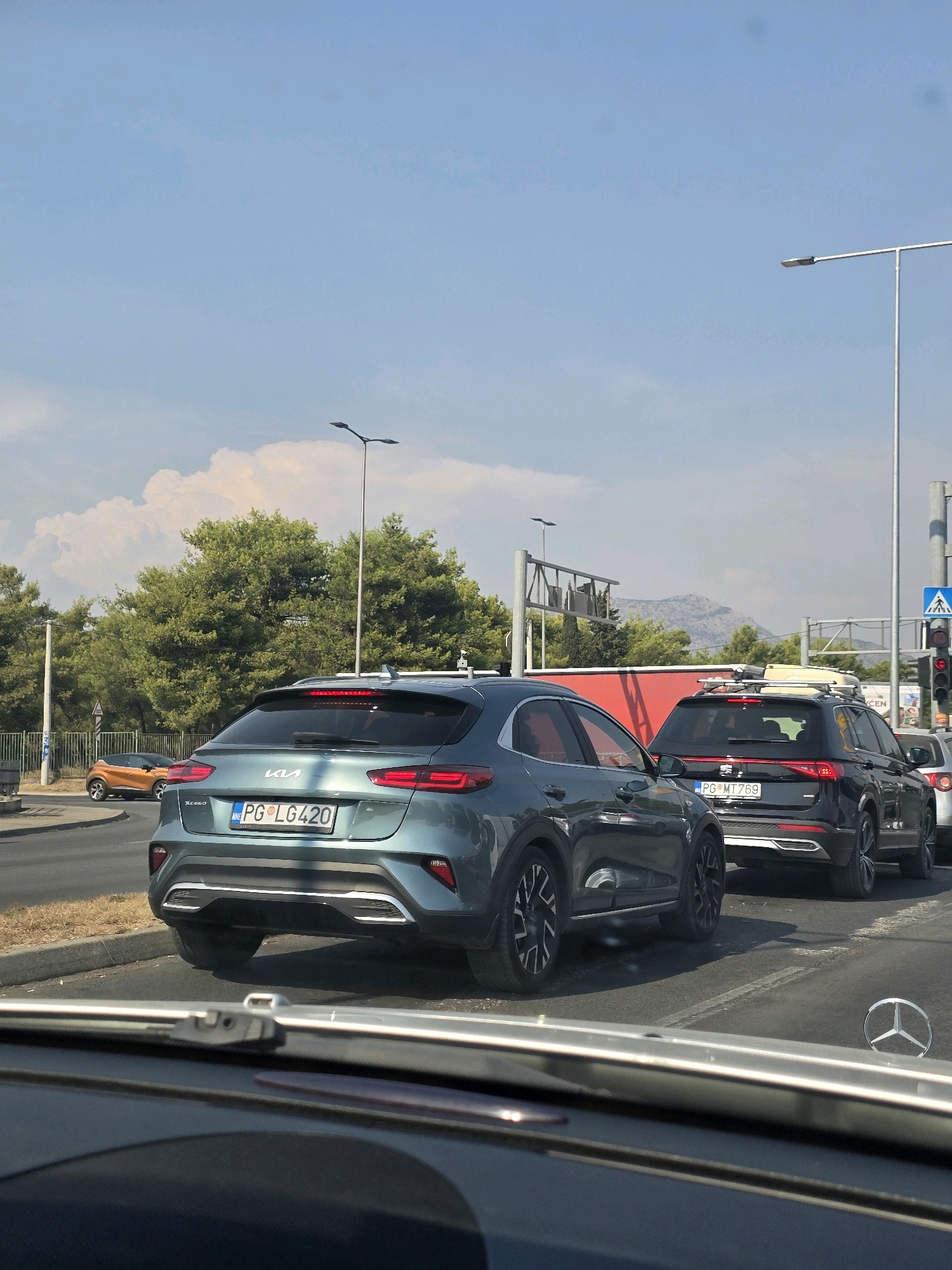 This image captures a traffic jam on a sunny day in Podgorica, Montenegro. Several cars are stopped on a multi-lane road lined with green trees and lampposts. The foreground shows the interior of a vehicle, including a portion of the dashboard and windshield wiper. In the middle ground, a teal Kia XCeed with license plate PG LG420 is prominently featured, followed by a black SUV with a roof rack and license plate PG MT769. An orange smaller car is visible further ahead on the left. In the background, mountains can be seen through the trees under a bright blue sky with scattered clouds. The overall scene suggests a typical urban traffic situation with cars waiting at a red light or due to congestion.