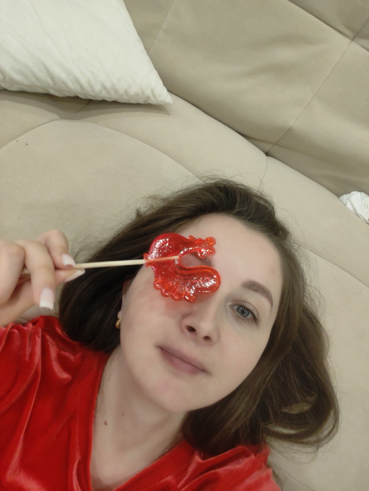 A selfie of a woman, wearing a red velvet shirt, lying on a beige couch next to a white pillow. She holds a red, heart-shaped lollipop on a stick in front of her face, partially covering one eye. The background is a neutral-toned couch and a partial glimpse of a white object.