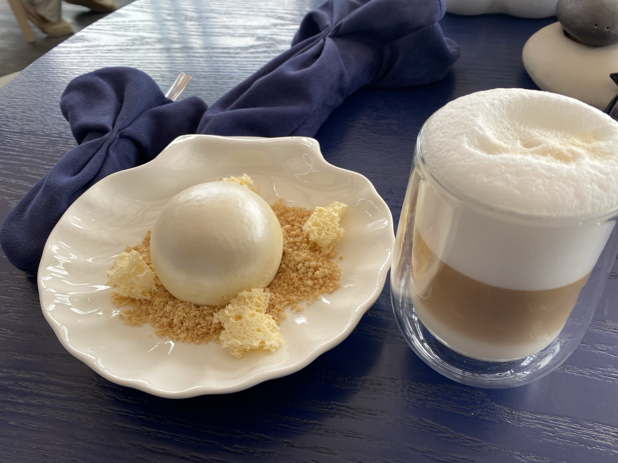 A top-down shot shows a dessert and a beverage on a blue, possibly lacquered, table. The dessert features a smooth, round, white dome on a decorative, scallop-edged plate. It is surrounded by a light brown crumbly base and irregular pieces of a porous, light yellow substance. Next to the dessert is a clear glass containing a layered drink: a light brown liquid at the bottom, topped with a thick layer of white foam. The environment includes a folded navy blue napkin and a glimpse of another object in the background. There is no indication of the city of Yekaterinburg or any people in the image.