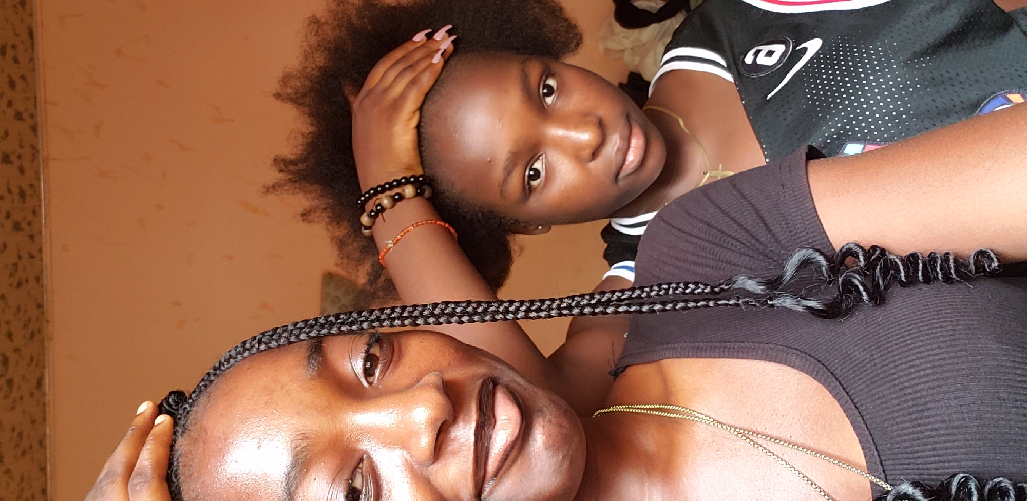 The image, captured indoors in Rido, Nigeria, features two young Black females in a close-up, likely selfie-style portrait.
The individual on the left, partially framed from the chest up, has dark skin and an apparent age in her late teens or early twenties. She wears her dark hair in box braids, with some individual braids featuring curly ends that extend downwards. Her left eye, nose, and a smiling mouth with a faint hint of her tongue are visible. She wears a black ribbed top and a thin gold chain necklace. Her right hand, with manicured nails, is placed gently on the head of the second individual.
The second individual, also appearing in her teens, is positioned slightly behind and to the right, mostly visible from the shoulders up. She has natural, voluminous dark afro-textured hair and clear skin. She looks directly at the camera with a neutral to slight smile. She wears a black athletic jersey with white striped trim on the collar and shoulders, and a white Nike logo with small dots on the chest. A gold cross pendant necklace is visible around her neck. On her right wrist (viewer's left), she wears multiple dark beaded bracelets and one thin orange beaded bracelet. Her fingernails are painted a light pink or nude color.
The background consists of a plain, light orange-brown wall with some subtle textural variations or minor blemishes. The lighting is soft and even, illuminating the subjects' faces.
