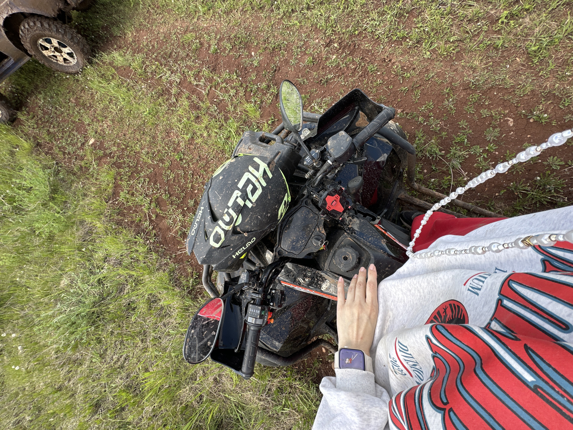 The image, possibly taken from an elevated viewpoint, depicts a black all-terrain vehicle (ATV) parked on a patch of land with a mix of dirt and sparse green vegetation. A black helmet with the word "HELMO" in a prominent yellow font rests on the ATV's front rack. A person wearing a white sweatshirt with red and blue graphics and a pearl necklace is positioned to the right of the ATV, with their left hand near the ATV's controls. The person is also wearing a smartwatch with a light-colored band. In the upper left corner, the tire of another vehicle is partially visible. The location is estimated to be Ilyinsky, Russia, due to the request within the prompt, though there are no other factors that point towards this.