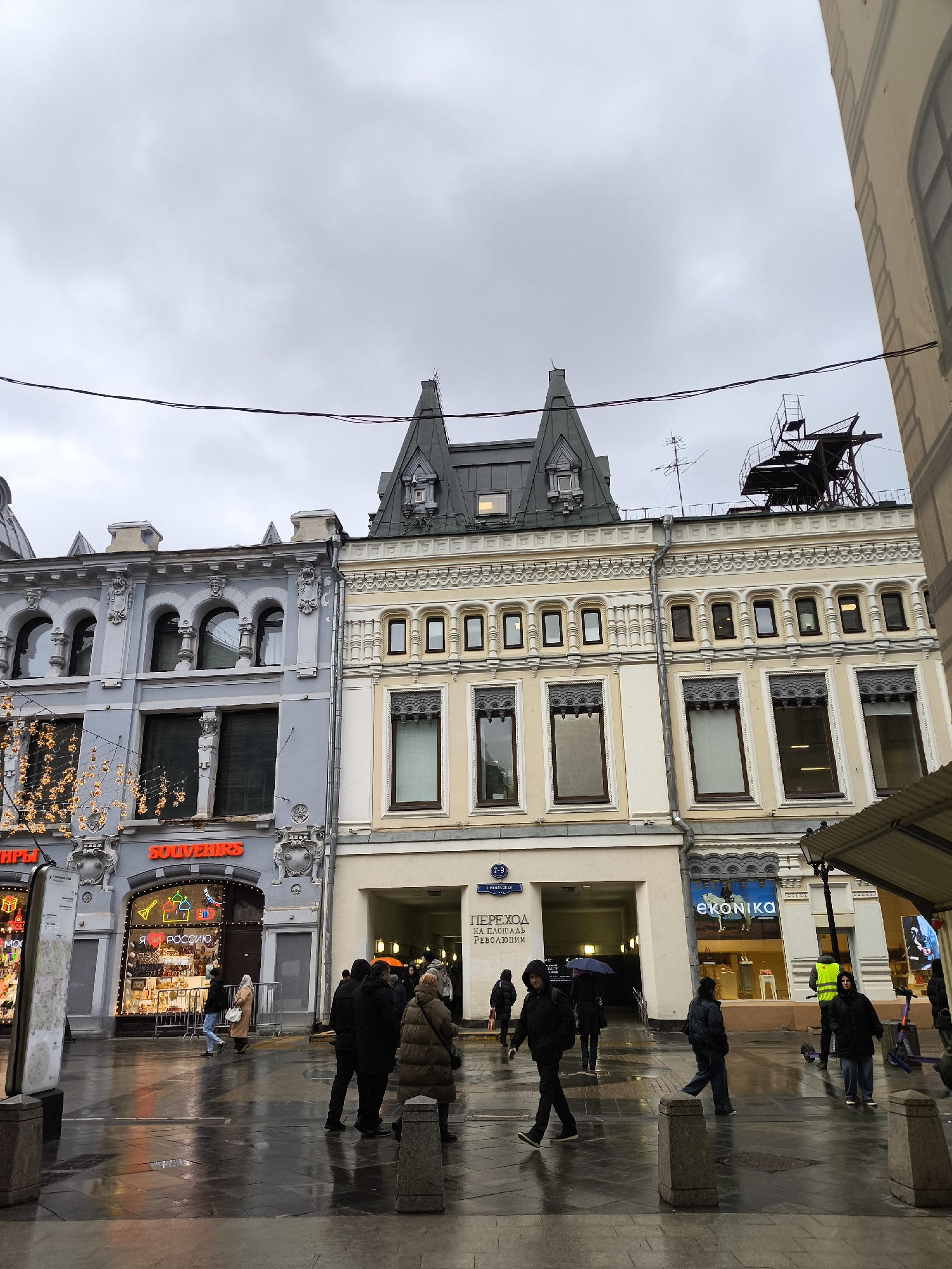 A street scene in an urban environment, likely in Russia based on the Cyrillic text on signage, captured on a cloudy, possibly rainy day.
The main subject consists of several buildings with distinct architectural styles. On the left, a light blue building is decorated with architectural elements and illuminated string lights. Adjacent to it, a yellow-beige building has arched windows and decorative trim. Between the two buildings is an archway that leads to an interior space, above which is a street address sign in Cyrillic. On the right side of the building, there's a store with a large window display featuring various shoes. On the roof of one of the buildings is what appears to be an iron staircase-like structure and an antenna.
People are walking on the wet pavement, some carrying umbrellas. They are dressed in warm clothing, suitable for the weather. The ground is wet, reflecting the streetlights and store signs.
The overall impression is a typical urban street scene in a European city, with a mix of commercial establishments and pedestrians navigating a potentially rainy or overcast day.