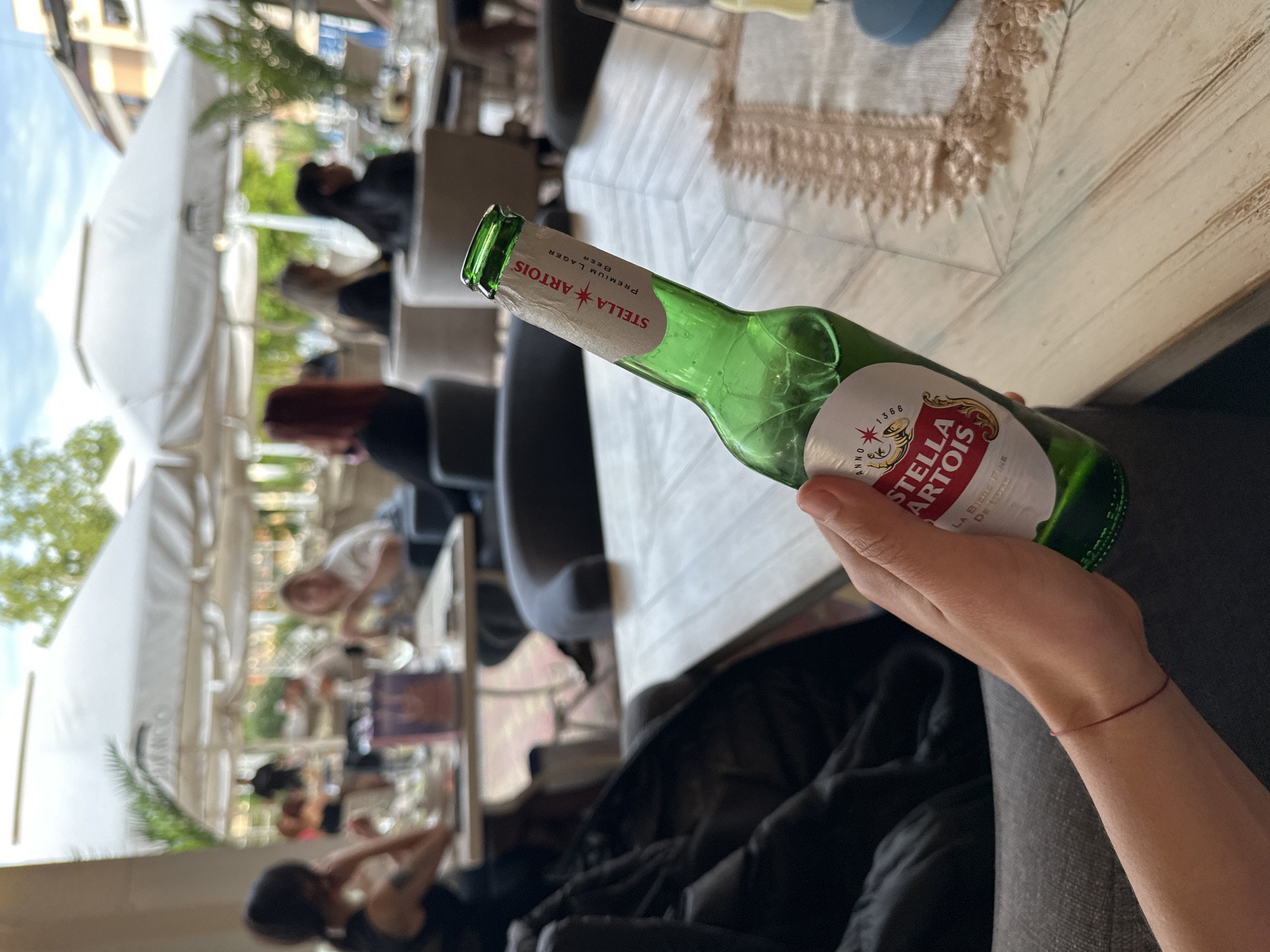 A slightly tilted, medium shot photograph depicts a person holding a partially empty bottle of Stella Artois beer in an outdoor setting, likely a café or restaurant. The bottle is predominantly green with the Stella Artois label visible, grasped by a hand with a thin red bracelet on the wrist. The background features a busy outdoor area, with other patrons sitting at tables under white canopies or umbrellas. The location appears to be in Veliko Tarnovo, Bulgaria, based on general architectural styles typical of such a historical region. Tables with textured white tablecloths are visible, and chairs are positioned around them. People are seen seated, engaged in conversation and other leisure activities. The lighting suggests it is daytime, with some areas illuminated brightly, and others in shadow. The overall atmosphere suggests a relaxed, social gathering.