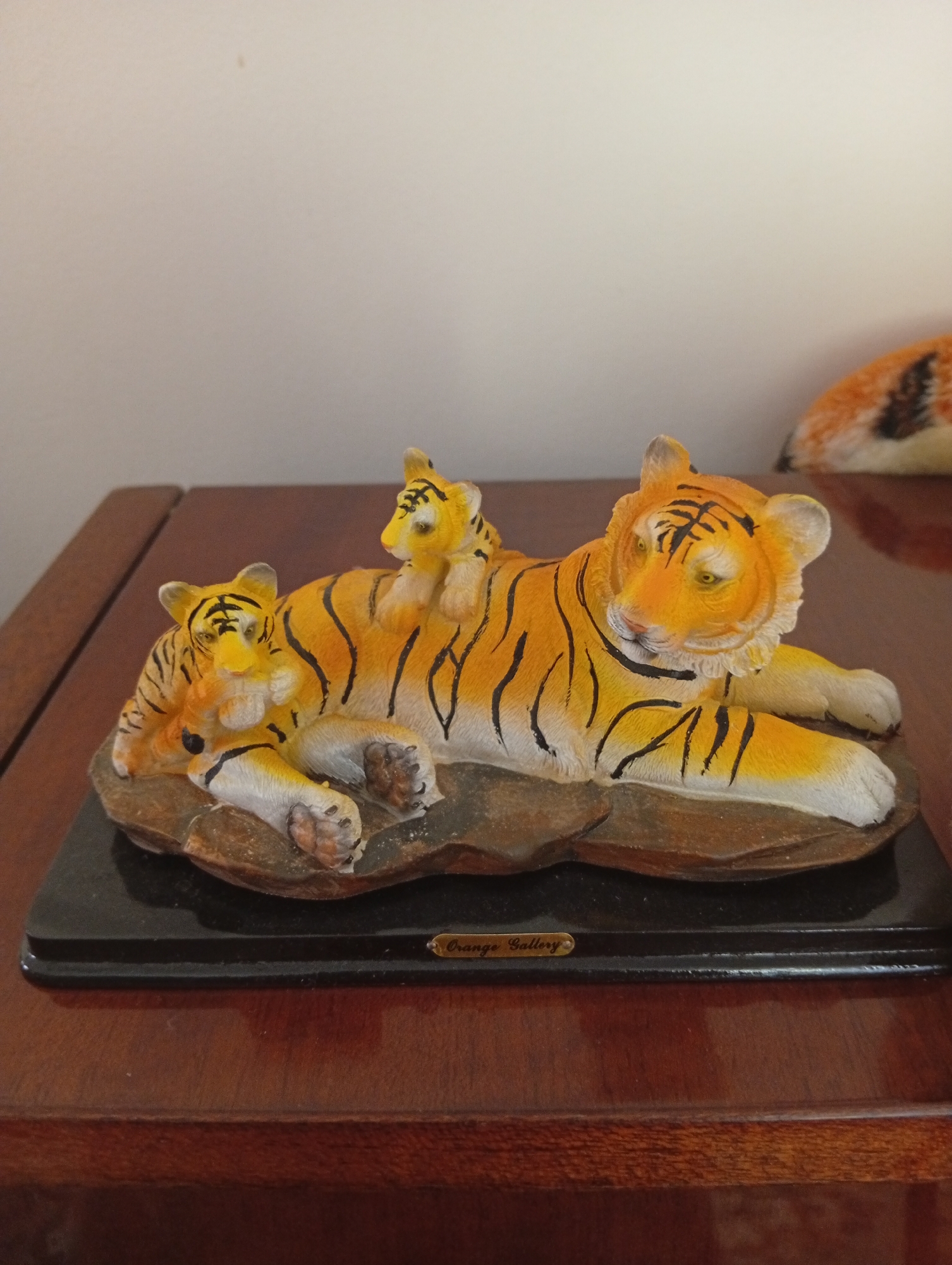 A tiger figurine with two cubs rests on a wooden surface in Velyatyno, Ukraine. The main figurine depicts an adult tiger lying down, with one cub nestled beside its head and another perched on its back. The tigers are orange and black, with detailed stripes and facial features. The figurine is mounted on a dark rectangular base, which is further placed on a polished wooden table. The background is a plain, light-colored wall. A small oval plaque on the base reads "Orange Gallery". The lighting suggests an indoor environment with diffused light from the side.