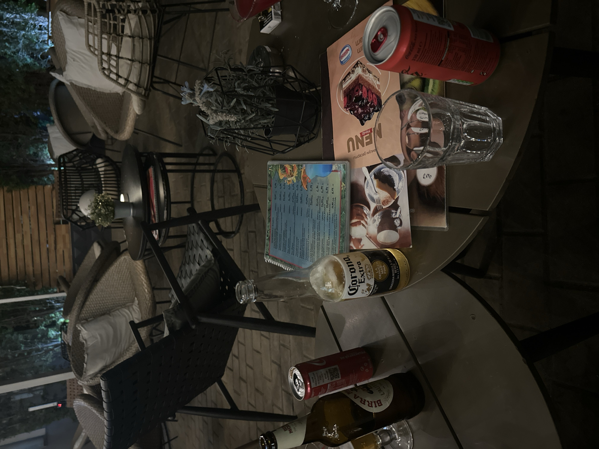 The scene depicts a table at an outdoor cafe, possibly in Pleven, Bulgaria, evidenced by the Cyrillic text on the menu. The table is cluttered with items suggesting recent use: a Coca-Cola can, an empty glass, a menu with a dessert image, and another menu displaying cocktail prices in leva. Additionally, there is an empty Corona beer bottle, another beer bottle labeled "BIRRA MORETTI," and an empty glass. A decorative plant in a wire container sits nearby. The surrounding environment shows outdoor seating with rattan chairs and the presence of trees, indicating an outdoor cafe setting. The lighting suggests it is evening.
