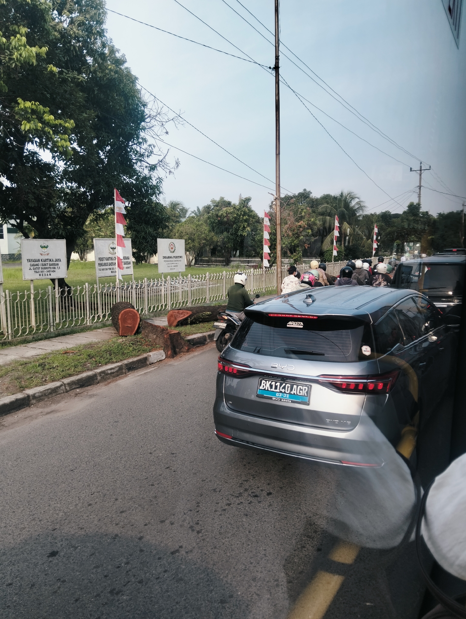 The image captures a street scene in Medan, Indonesia, likely near the Polsek Medan Kota or a similar official establishment, given the presence of official-looking signs and white fencing. A gray BYD Arista car, with license plate BK 1140 AGR, is the primary subject in the foreground, facing away from the camera and parked on the right side of a paved road. The car is positioned in traffic, with several motorcycles and other vehicles visible behind it.
On the left side of the road, a sidewalk is bordered by a white fence. Beyond the fence, three white signs with red and white Indonesian flags are displayed. The signs appear to be informative plaques, possibly related to local government or community organizations. A large, recently felled tree trunk and several large branches are lying on the grass next to the sidewalk, indicating recent tree removal.
A person wearing a white helmet is on a motorcycle to the left of the gray car, also facing away from the camera. Other individuals on motorcycles are visible further down the road, forming a line of traffic. The environment appears to be a well-maintained urban area with trees lining the street and a clear, pale sky overhead. Power lines are strung across the sky, supported by poles. The reflection of the surrounding environment can be seen on the car's windows, suggesting the photo was taken from inside another vehicle.