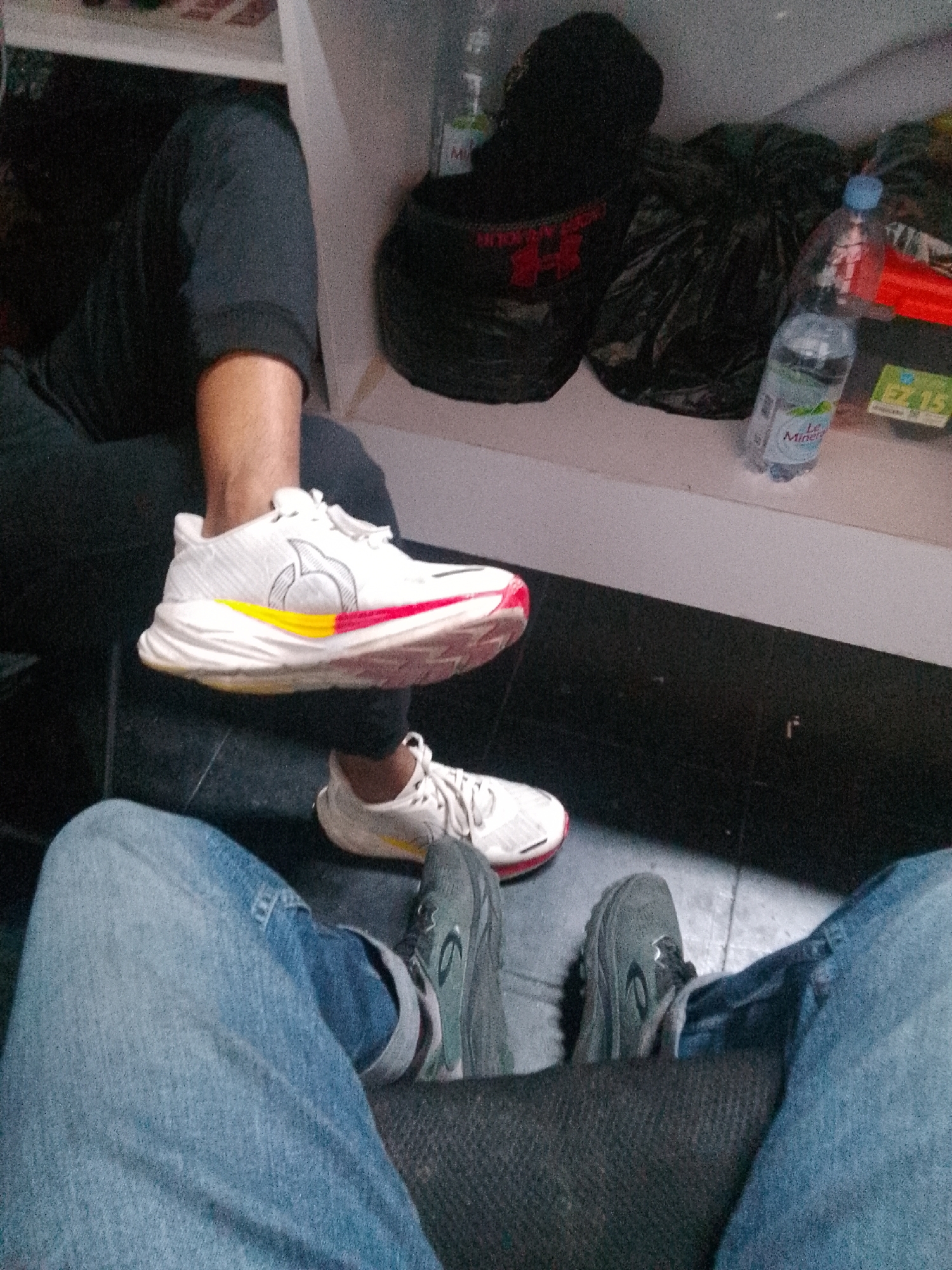 In Trenggalek, Indonesia, a person is sitting on a dark-colored textured seat. They are wearing blue jeans and dark grey athletic shoes. In front of them, another person's legs are visible. This individual is wearing black pants and white athletic shoes with red and yellow accents. The shoes appear to be branded with a circular logo. The background shows a white shelf with several items including black plastic bags, a clear plastic bottle of water labeled "Le Minerale," and other indistinguishable objects. The flooring is dark. The overall scene suggests an indoor environment, possibly a waiting area or casual resting spot.