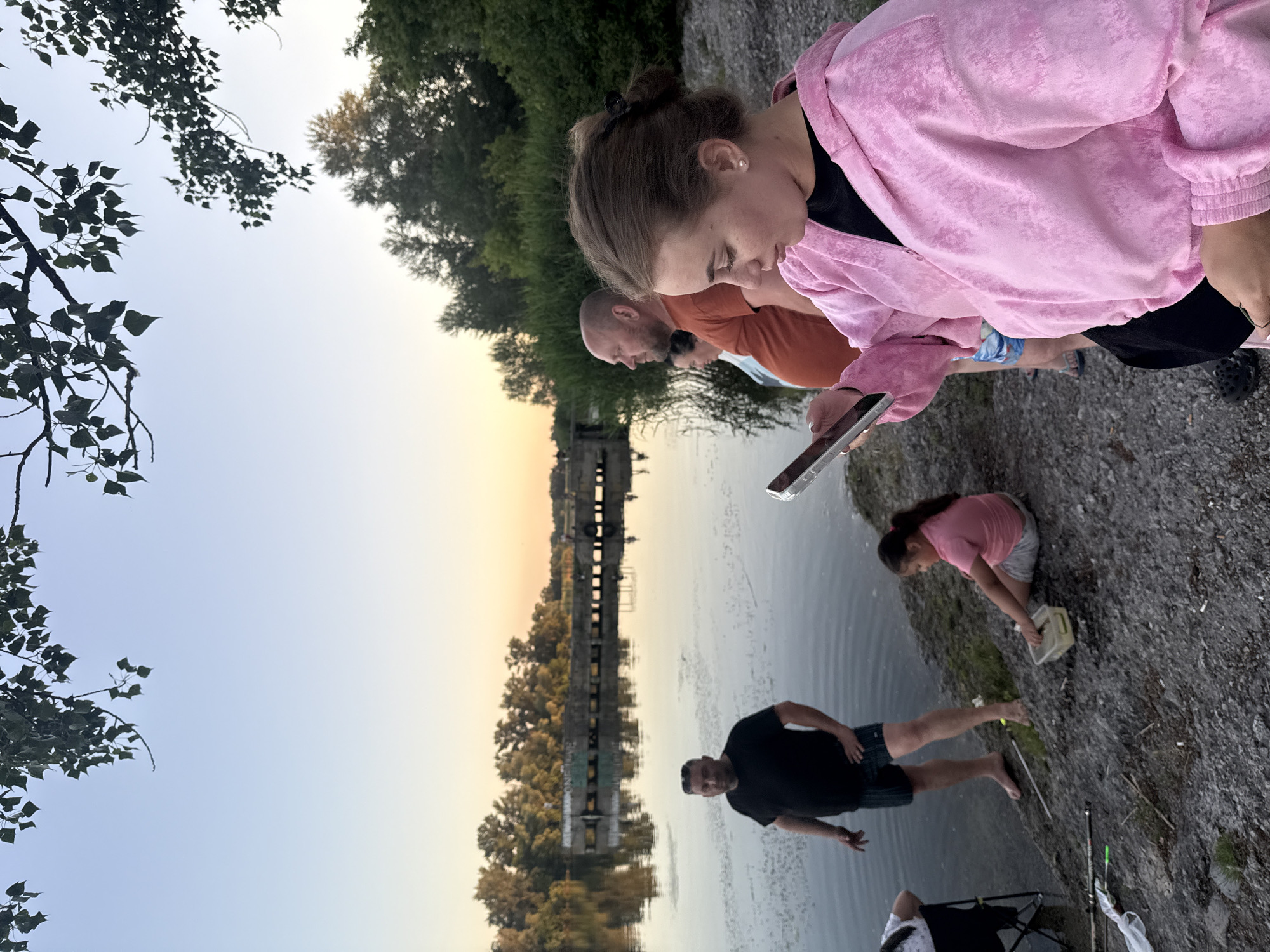 This image depicts a group of people by a body of water, likely a river or lake, with trees and a bridge structure in the background. The sky is clear with a soft gradient, suggesting it is either sunrise or sunset. The ground is rocky, typical of a riverbank.
On the right, a young woman in a pink velour hooded jacket is looking at a tablet. Next to her, a man in an orange t-shirt and checkered shorts stands with his arm extended. Behind him, another person is partially visible.
To the left, a man in a black t-shirt and dark shorts stands near the water's edge, looking out at the water. A young girl in a pink t-shirt and grey shorts is sitting on the bank, holding a small plastic container. To the far left, a person is seated in a chair, partially visible. Fishing rods and tackle are scattered on the ground near the water. The bridge structure in the background appears to be an old, possibly disused, concrete dam or industrial facility, with graffiti visible on its surface. The water reflects the trees and the sky.
This scene is consistent with the banks of the Dnipro River in the city of Dnipro, Ukraine, particularly in areas with remnants of industrial infrastructure and natural shorelines.