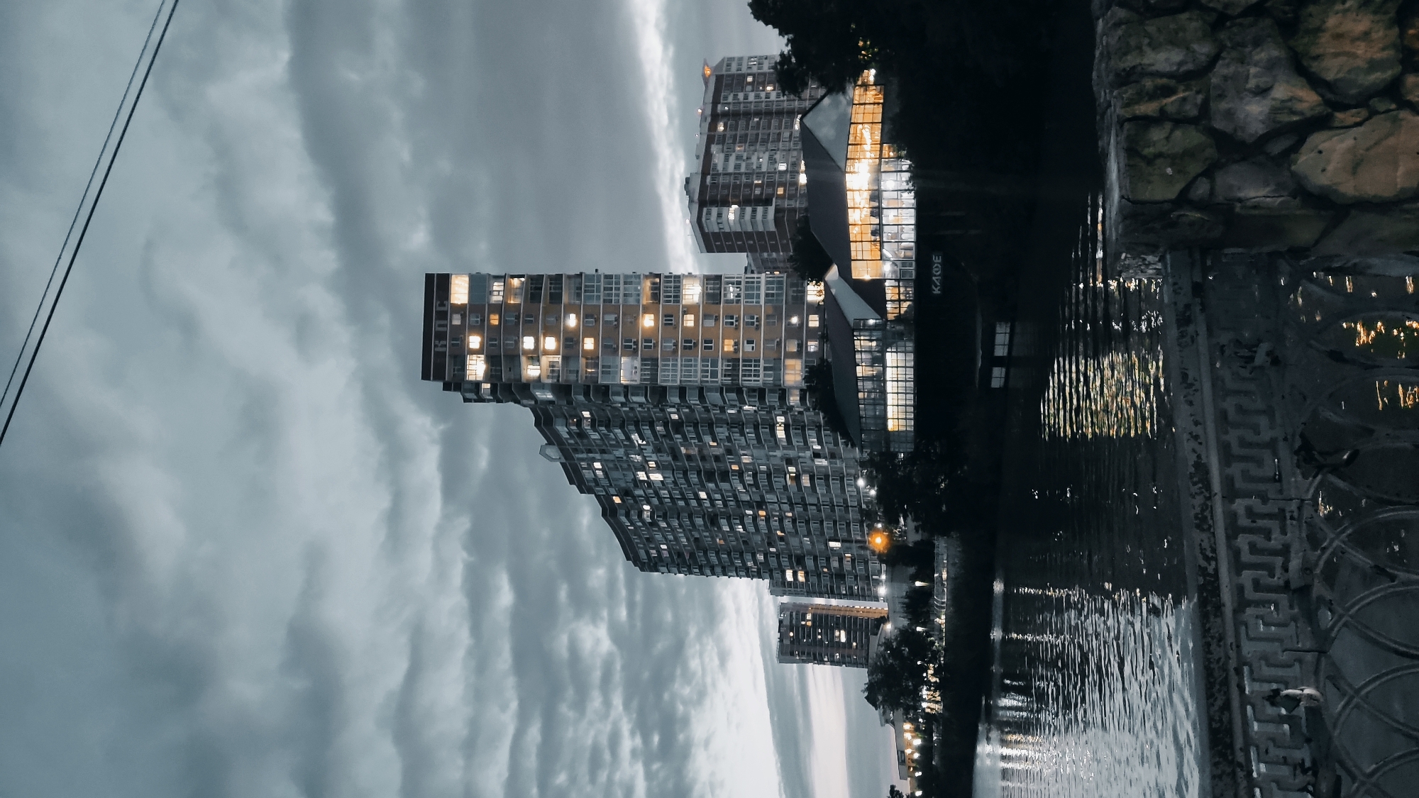 The image depicts a view of the city of Krasnodar, Russia, at dusk. A body of water, likely a river or canal, occupies the foreground, reflecting the lights of buildings and the sky. The water is calm, with ripples disturbing the reflections. A decorative stone and metal railing runs along the edge of the water in the lower portion of the frame.
Dominating the background are several large, modern apartment buildings. The tallest building on the left has an unusual, stepped silhouette and is illuminated by many internal lights, suggesting it is inhabited. To its right, another, slightly shorter apartment building also shows illuminated windows. In front of these buildings, a low-rise structure with extensive glass walls is visible, with warm lights emanating from within, indicating it may be a restaurant or cafe, as suggested by the Cyrillic word "КАФЕ" (KAFE) visible on its facade. Trees and dark foliage line the bank between the water and the buildings, obscuring some of the lower levels. The sky is overcast with dramatic, dark clouds, contributing to the moody, twilight atmosphere. Power lines are visible stretching across the top of the frame. No people are visible in the image.