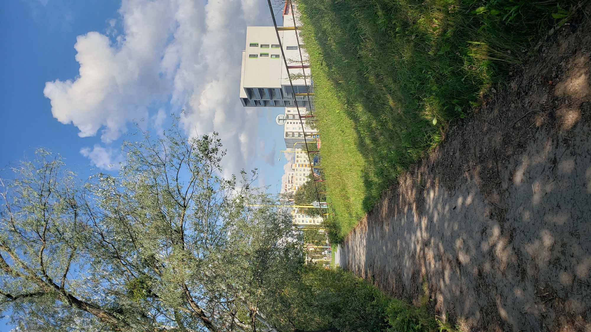 This image depicts a view of a pathway next to a grassy embankment, with a residential area and apartment buildings in the background. The pathway appears to be made of dirt and gravel, with fallen leaves scattered on its surface, suggesting it is an outdoor, possibly park-like setting within the city. The grassy embankment is covered in lush green grass, with some weeds and small plants growing amongst it. A metal fence, painted with red, yellow, and white stripes, runs along the top of the embankment.
In the background, several apartment buildings are visible. The closest building is a modern, multi-story structure with a white facade and green-framed windows. Further in the distance, more apartment buildings of varying architectural styles can be seen, some appearing to be older, block-style structures common in Eastern European cities. The sky above is a bright blue with scattered white clouds, indicating fair weather.
The image captures a scene in the city of Prešov, Slovakia, specifically in the Sekčov housing estate. The pathway is likely a pedestrian path or part of a walking trail within this urban environment, connecting to or passing by residential areas. The presence of trees and vegetation alongside the pathway suggests an effort to integrate green spaces within the city. No people are visible in the image, and no specific actions or interactions are taking place. The overall impression is one of a quiet, everyday urban landscape on a clear day.