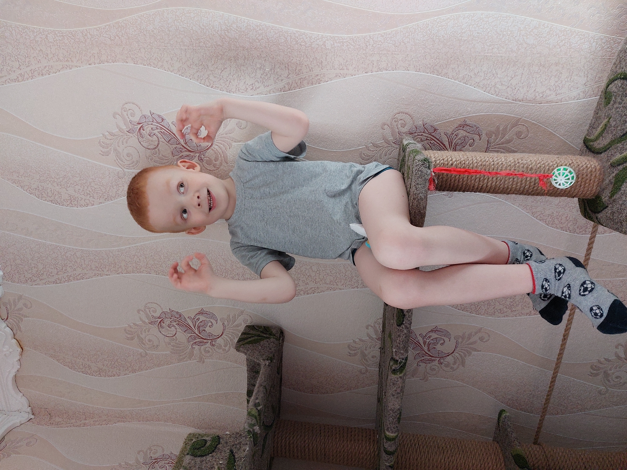 A red-haired child wearing a gray t-shirt and socks with soccer ball patterns is lying on a cat tree in front of a wallpapered wall, in Большой Букор, Russia. The child is smiling and holding his hands in a circular gesture. The cat tree is made of carpeted posts and rope.