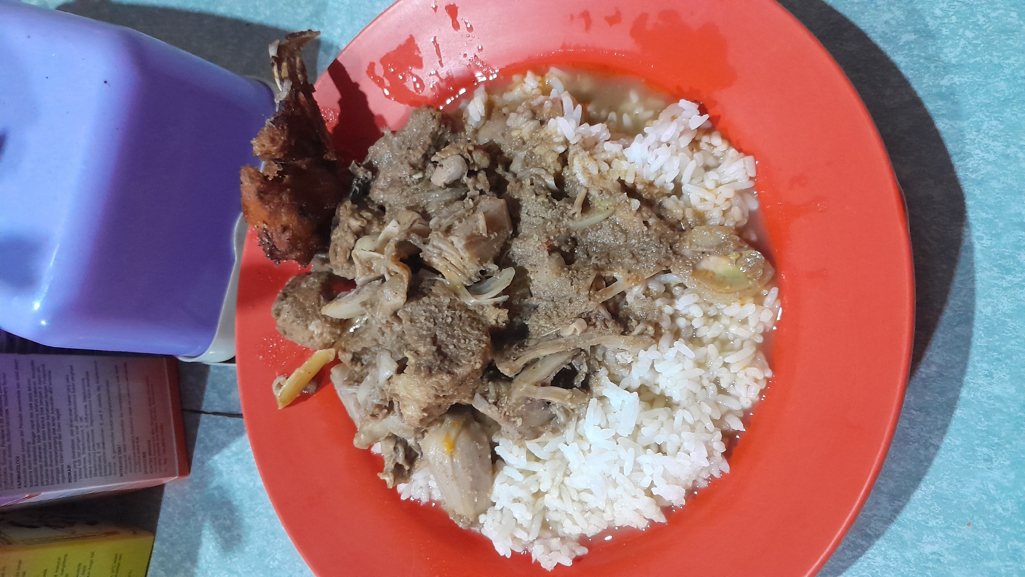 A red plate holds a portion of white rice covered with a meat dish, likely beef or goat, and a fried piece of meat to the left of the rice. The plate sits on a light blue surface, possibly a table. A light purple container and boxes of medicine are present in the lower left of the frame. The lighting appears to be artificial, casting distinct shadows. The location is likely a restaurant or home in Lhokjok, Indonesia, based on the food served.