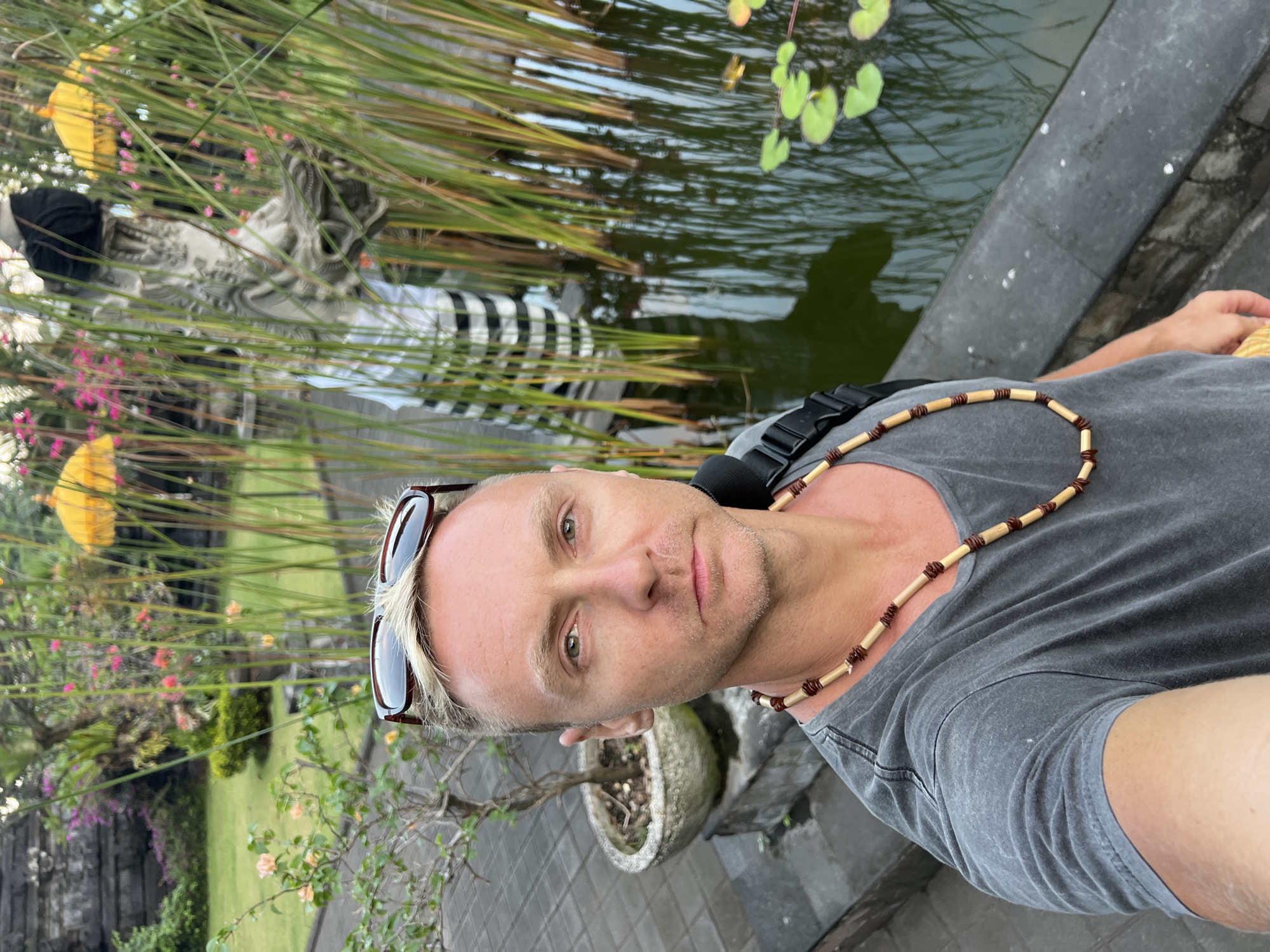 A man with blond hair and sunglasses on his head, wearing a gray t-shirt and a beaded necklace, takes a selfie in front of a pond with reeds and lily pads. In the background, there is a stone statue dressed in a traditional black and white striped sarong, and two yellow umbrellas. The location is a temple in Dawan, Indonesia.