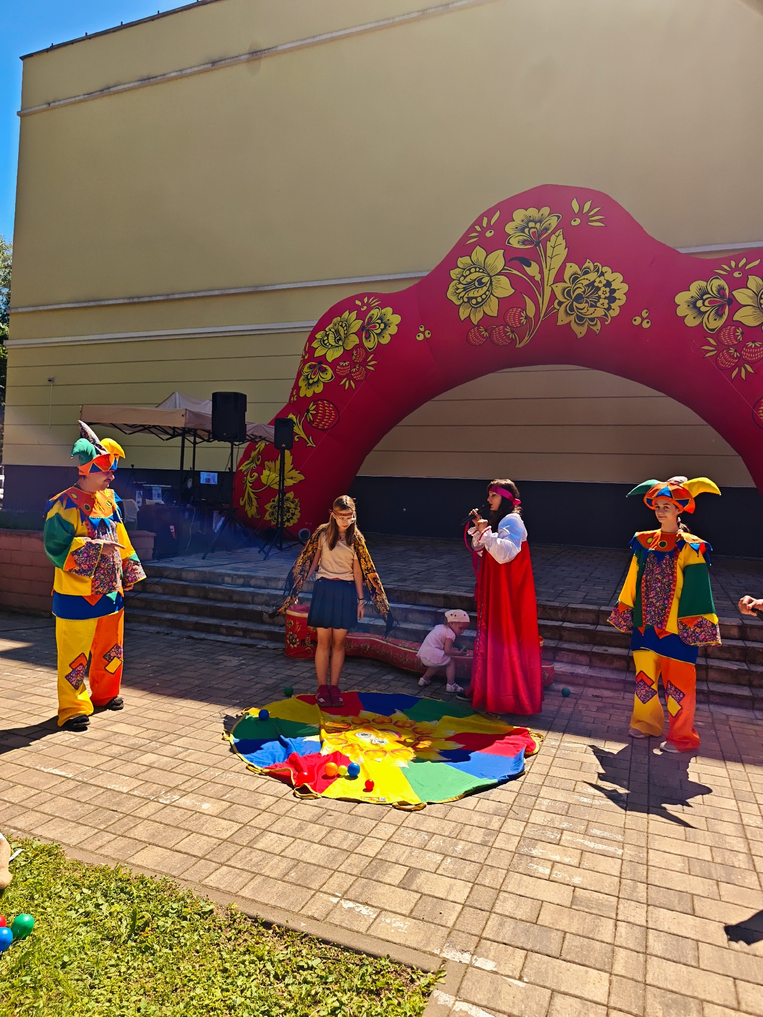 This image depicts an outdoor public event taking place in Dubna, Russia. Five individuals are present: four adults and one small child.
In the left portion of the foreground, an adult male dressed in a vibrant, multi-colored harlequin-style costume with a pointed hat and sunglasses stands, pointing forward with his right hand. Directly to his right, a young adult female wearing a brown top, dark skirt, and a patterned shawl around her shoulders stands on a circular, multi-colored mat (featuring red, blue, green, and yellow sections) laid on the light-colored paved ground. She has a white blindfold or mask covering her eyes. Scattered on and around the mat are several small colored balls.
To the right of the blindfolded female, an adult female in a long red dress with a white undershirt and a red headband holds a microphone to her mouth, appearing to address the audience or lead an activity. Further to the right, another adult female in a multi-colored harlequin-style costume, similar to the male on the left, stands observing. Behind the woman in red, on a set of light-colored steps, a small child in a white outfit and cap is visible, seemingly crawling or playing.
The background is dominated by a large, red inflatable arch decorated with yellow floral and strawberry patterns, suggesting a traditional Russian Khokhloma art style. This arch functions as a decorative backdrop or entrance. Behind the performers and the steps, a pale yellow building with two horizontal white trim lines is visible under a clear blue sky. On the steps, a white canopy tent, two black speakers on stands, and additional sound equipment are arranged. The ground is paved with rectangular light-colored pavers, and green grass is present in the lower-left corner. The scene is brightly lit by strong sunlight, casting distinct shadows.