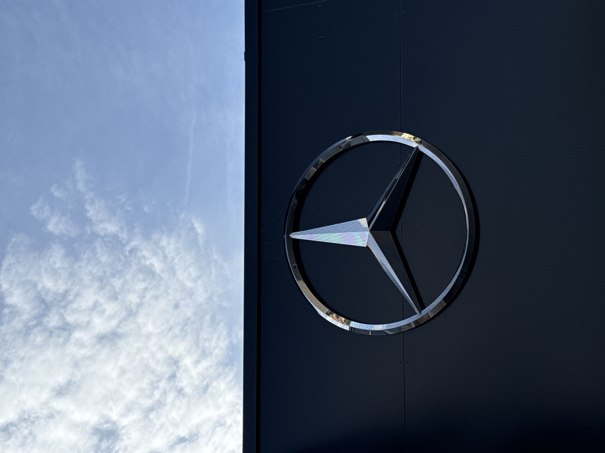 The image shows a silver Mercedes-Benz logo affixed to a dark blue/black building facade, possibly a dealership or service center. The logo is prominently displayed on the right side of the frame. To the left of the building facade, a portion of the sky is visible, featuring a light blue hue with scattered white clouds. The scene suggests an outdoor setting in Riedlingen, Germany, where the Mercedes-Benz building is likely located.