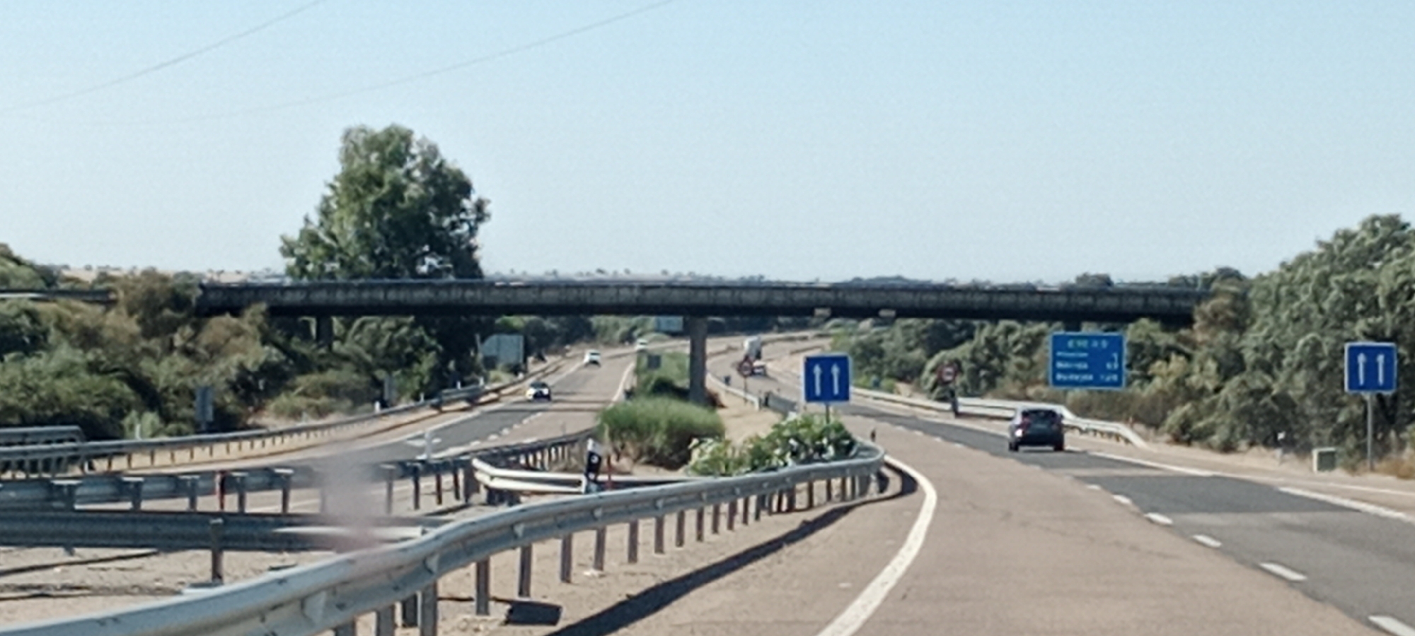 The image shows a highway overpass in Puerto de Santa Cruz, Spain. The sky is clear and blue. The highway has multiple lanes and is bordered by guardrails and vegetation. There are several cars on the road, including a black car and a white car. A large truck is also visible in the distance. There are several road signs, including a blue sign with white arrows indicating two lanes going in the same direction and another blue sign with text indicating destinations and distances. The overpass is a concrete structure with supports visible on either side of the road. The overall scene is typical of a highway in a rural or suburban area.