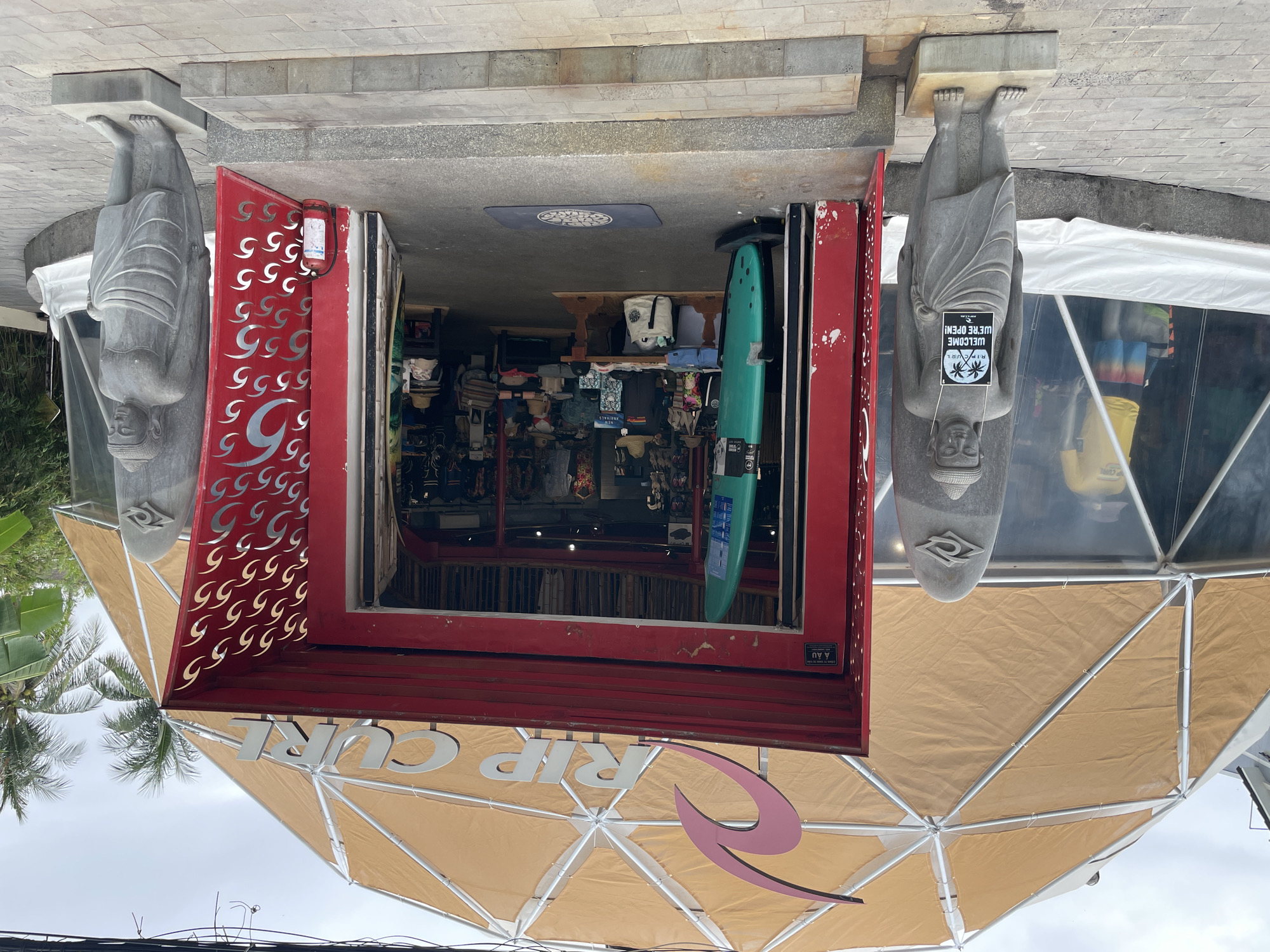 This is a shopfront of a Rip Curl store in Phu Thuy Ward, Vietnam. The store has a red-framed entrance with a geometric pattern on one side and is flanked by two stone statues that are holding signs that read "WELCOME WE'RE OPEN!" A surfboard is visible inside the store. Above the entrance is a beige and white dome-shaped structure with the Rip Curl logo. The sky is overcast, and palm trees can be seen to the left of the frame.