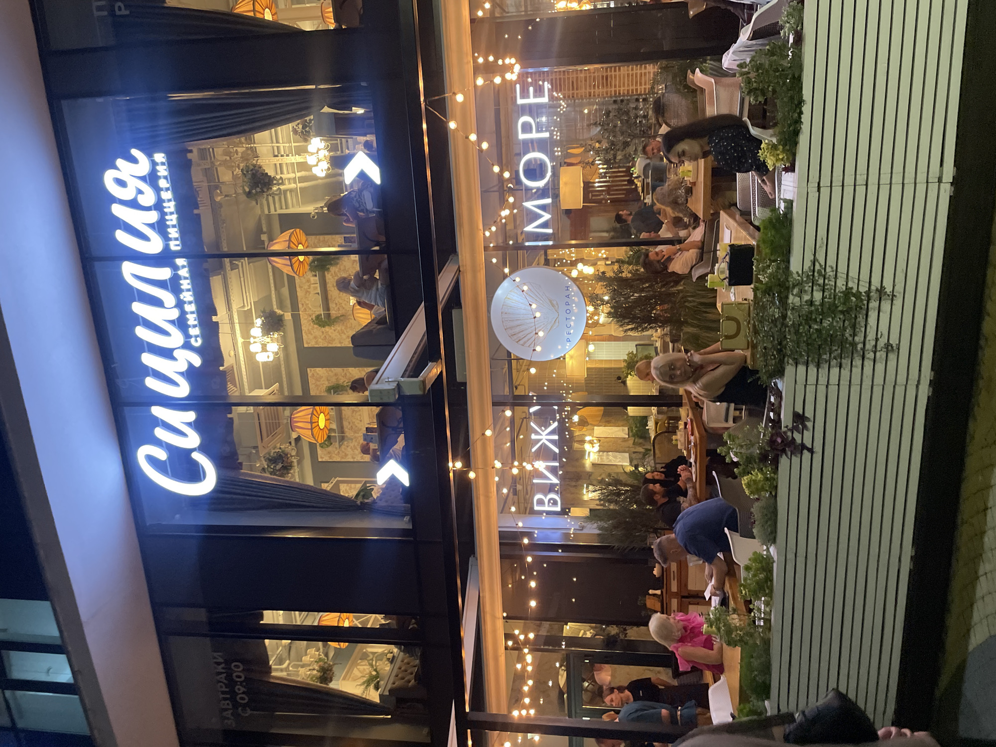 The image shows the exterior of a restaurant at night, with a brightly lit facade featuring large windows and illuminated signage. The restaurant's name appears to be "СИЦИЛИЯ" (Sicily) in large white illuminated letters, with smaller text below that reads "СЕМЕЙНАЯ ПИЦЦЕРИЯ" (Family Pizzeria). Through the windows, patrons can be seen dining and socializing at tables. String lights are visible on the exterior and within the restaurant.
Another sign visible on the lower level reads "IMORE" in illuminated letters, and to its left, the word "ВИЖУ" (I see) is also illuminated. Below the "IMORE" sign, a circular logo featuring a seashell and the word "РЕСТОРАН" (Restaurant) is visible.
Several people are visible in the foreground and through the windows. In the foreground, on a covered outdoor seating area, people are seated at tables, some appearing to be conversing and eating. The outdoor area is lined with white planters containing greenery.
The context provided indicates this is the restaurant "Sicily" in Novorossiysk, Russia. The facade suggests a modern, upscale dining establishment. The overall scene captures a lively evening at a restaurant.