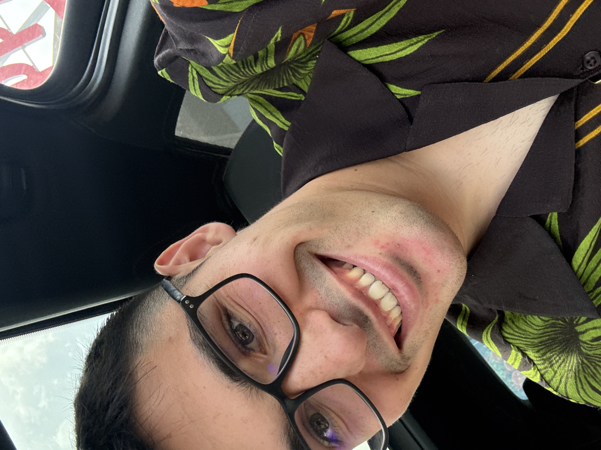 The image captures a close-up, low-angle shot of a young man smiling broadly from within what appears to be a car interior. He has short dark hair with a faded style and is wearing black-rimmed glasses that reflect a subtle purple-blue light. His wide smile reveals his teeth, with some natural yellowing visible on the lower incisors. He has light stubble on his chin and cheeks, and some minor skin redness or blemishes are present.
The man is dressed in a dark-colored, possibly black, collared shirt adorned with a vibrant tropical pattern of green leaves and yellow-orange stripes. Parts of the shirt's collar and an open button are visible around his neck.
The background elements indicate a car interior, with a dark headliner or roof lining visible on the left and top. Through a window in the upper left corner, a bright, overcast sky is discernible, along with a partial red object with white outlines, possibly a letter 'R' or part of a sign. The overall lighting suggests it is daytime. The man's joyful expression is the central focus. The image's context is Radinovo, Bulgaria.