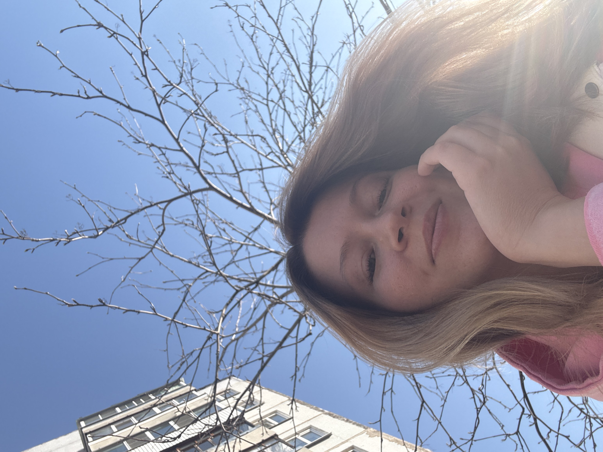 The image shows a woman with light brown hair in the foreground. She is wearing a pink hooded jacket and has her hand near her face. Behind her, a bare tree branch extends across a clear blue sky. In the lower left corner of the frame, a multi-story building with multiple windows is visible. The photograph appears to have been taken from a low angle, looking up. While the image may have been taken in Chaikovsky, Russia, there is no way to independently determine that location from the visible content of the image.