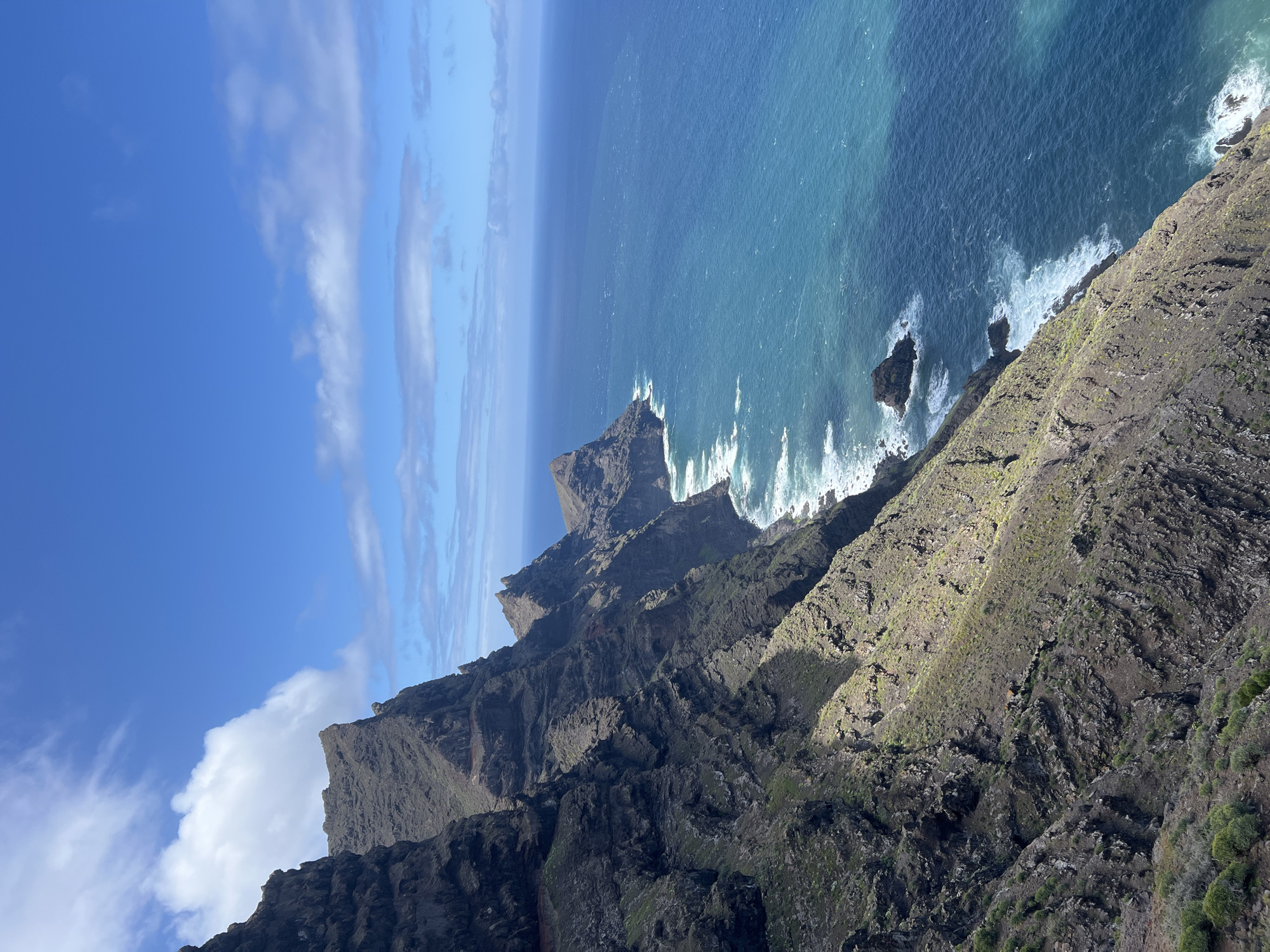 A high-angle view captures the dramatic coastline of Tirma, Spain. Steep, dark rocky cliffs covered with sparse vegetation plunge into the turquoise ocean, where waves crash against the base. A clear blue sky with scattered clouds is visible.